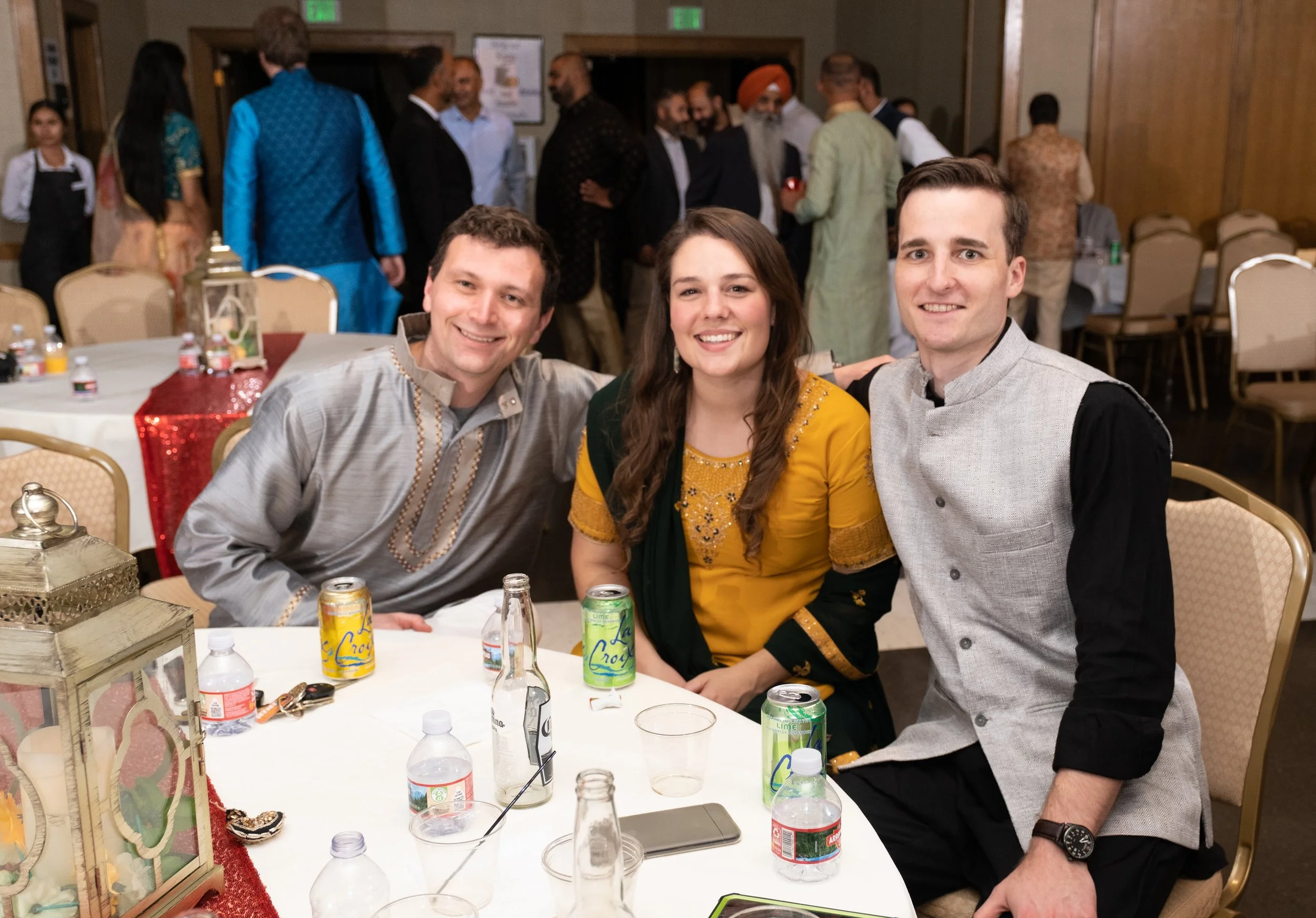 Three people sitting at a table smiling for the camera during a celebration. The table has various drinks, including soda cans and bottled water, and decorative items. In the background, a group of people are mingling, some dressed in traditional Ind