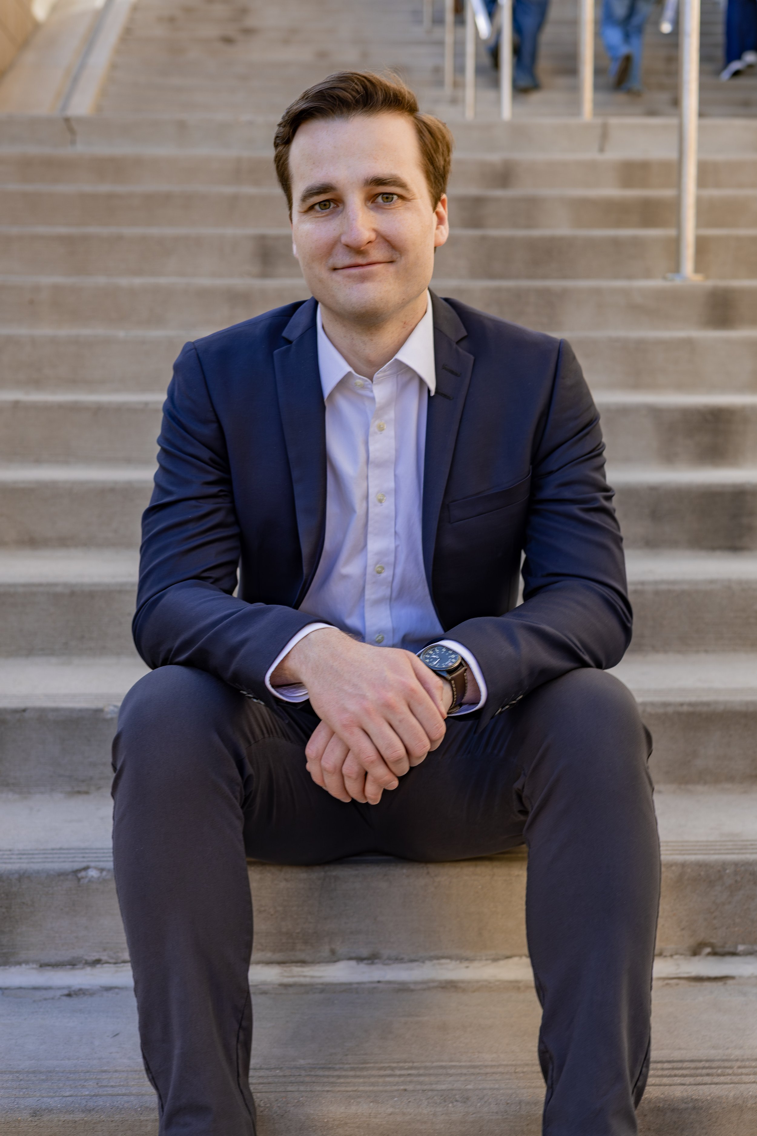 A young man in a navy blazer and white shirt sitting on stairs with a smile, hands clasped, wearing a watch, outdoors with people walking in the background.