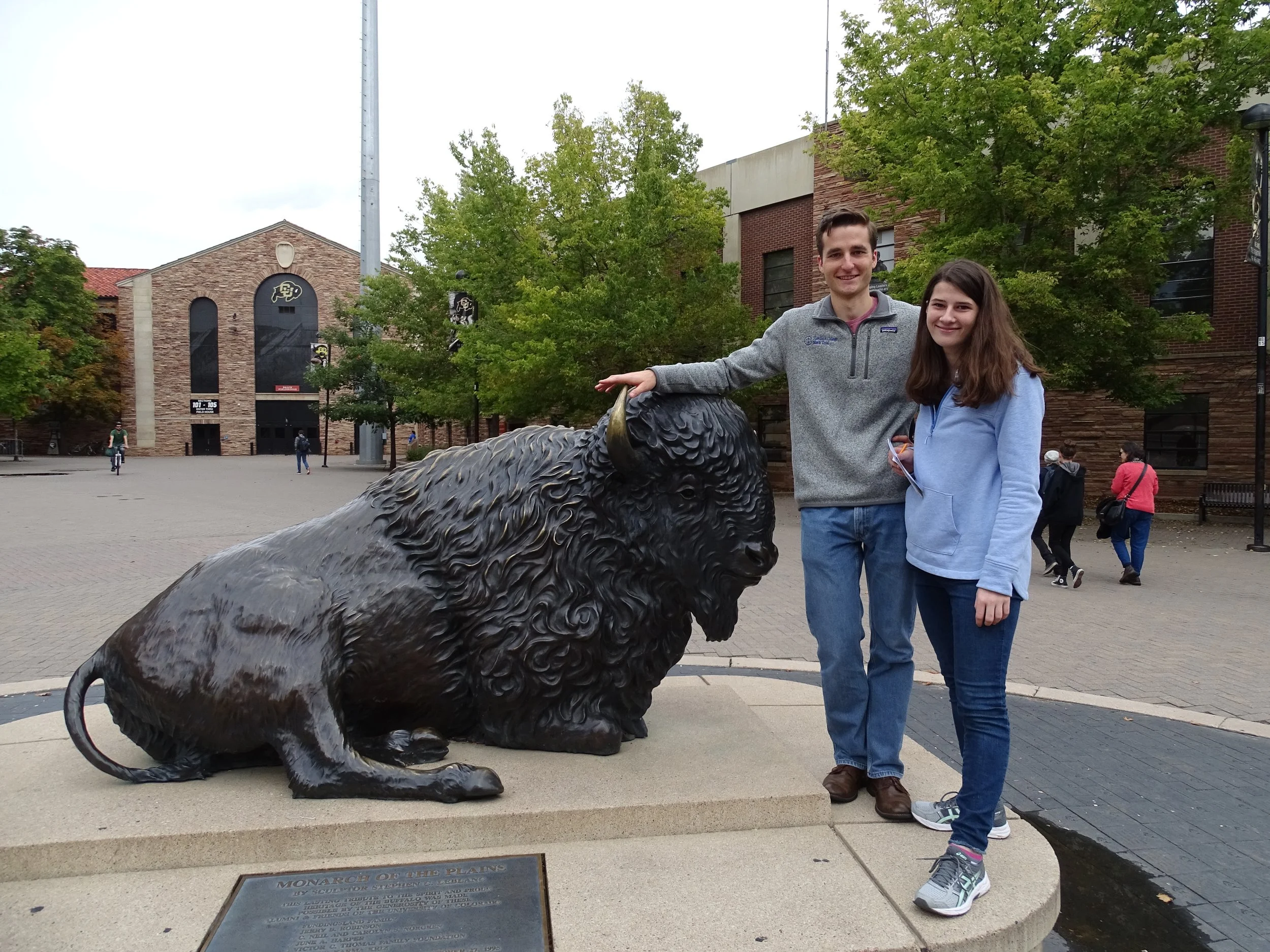 Two people standing next to a large bison statue in an outdoor plaza with trees and brick buildings in the background.