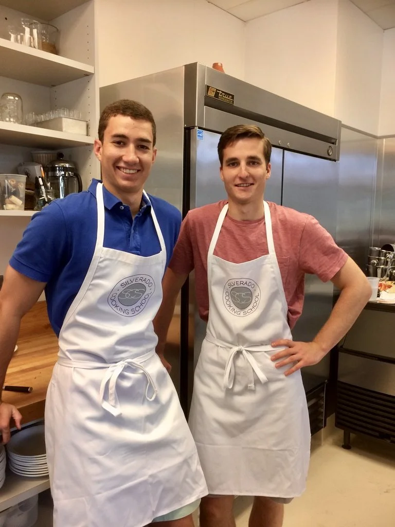 Two young men smiling and wearing white aprons with the Silverado Cooking School logo inside a kitchen with stainless steel appliances and shelves filled with kitchen supplies.