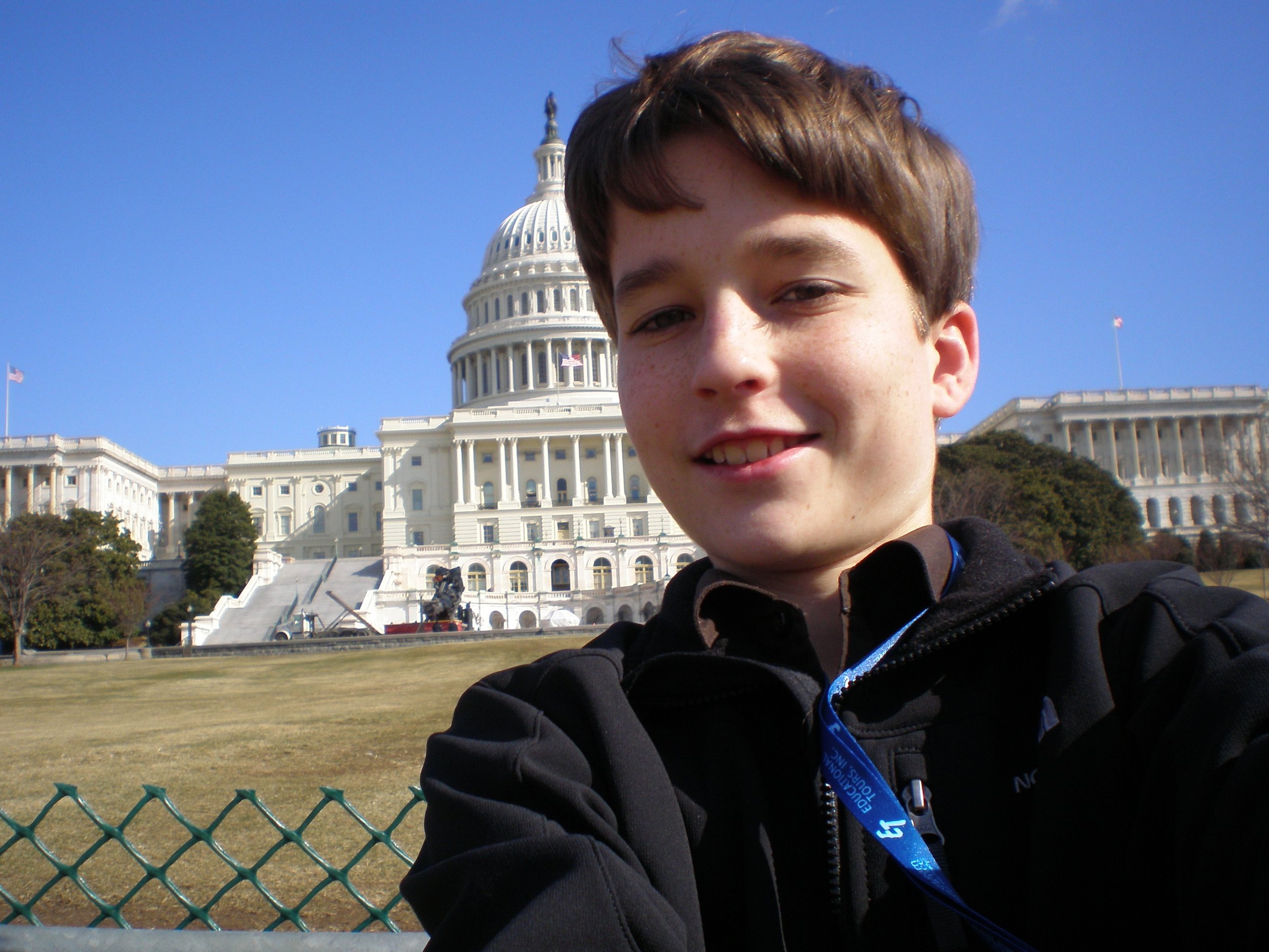 Smiling young boy in black jacket taking a selfie in front of the United States Capitol building on a clear day.