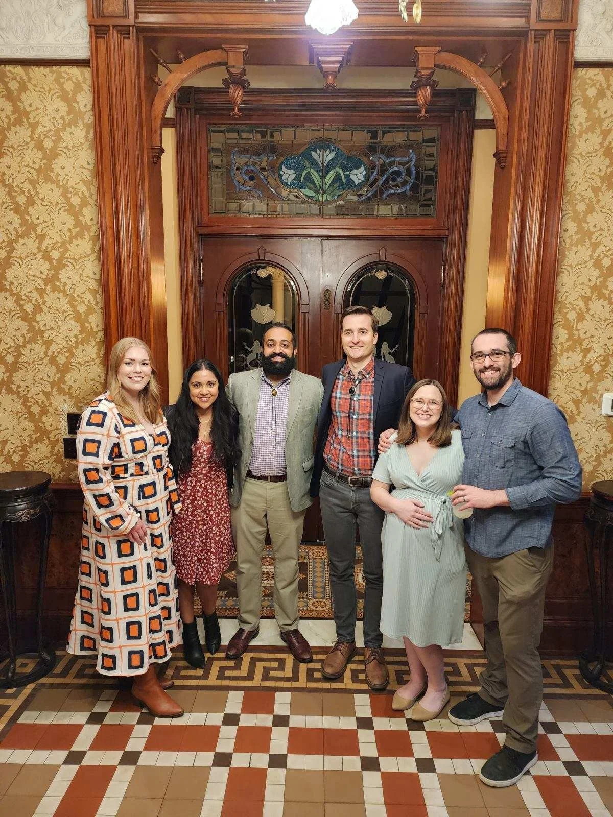 A group of six people standing together in a warmly decorated room with wooden trim and patterned wallpaper. They are smiling and posing for the photo, with a wooden door and stained glass window in the background.