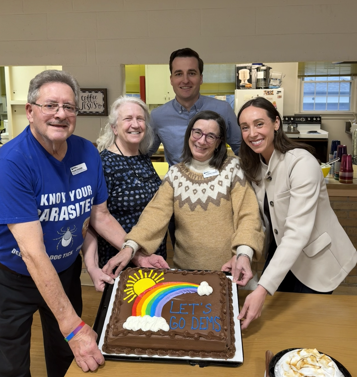 Group of five people gathered around a birthday cake decorated with a rainbow, sun, and clouds, with the text "Let's Go Dems" written on it, in a kitchen or community room setting.