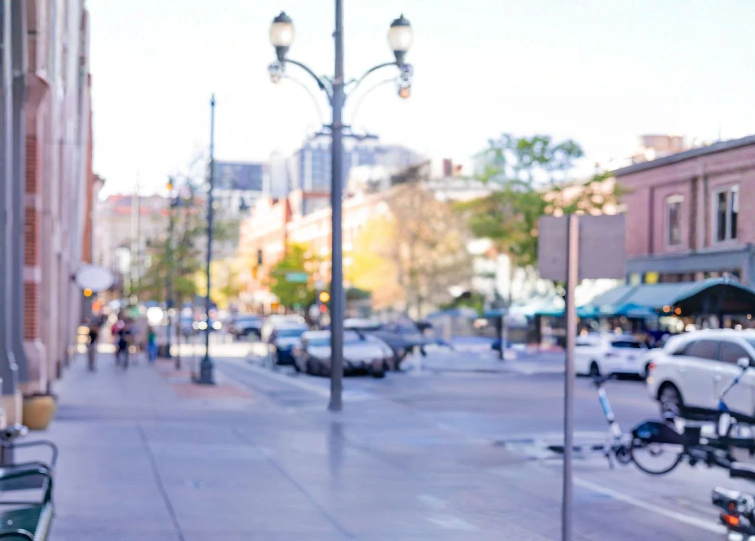 A blurry outdoor city street scene showing parked cars, sidewalk, lamp posts, and buildings with trees in the background.