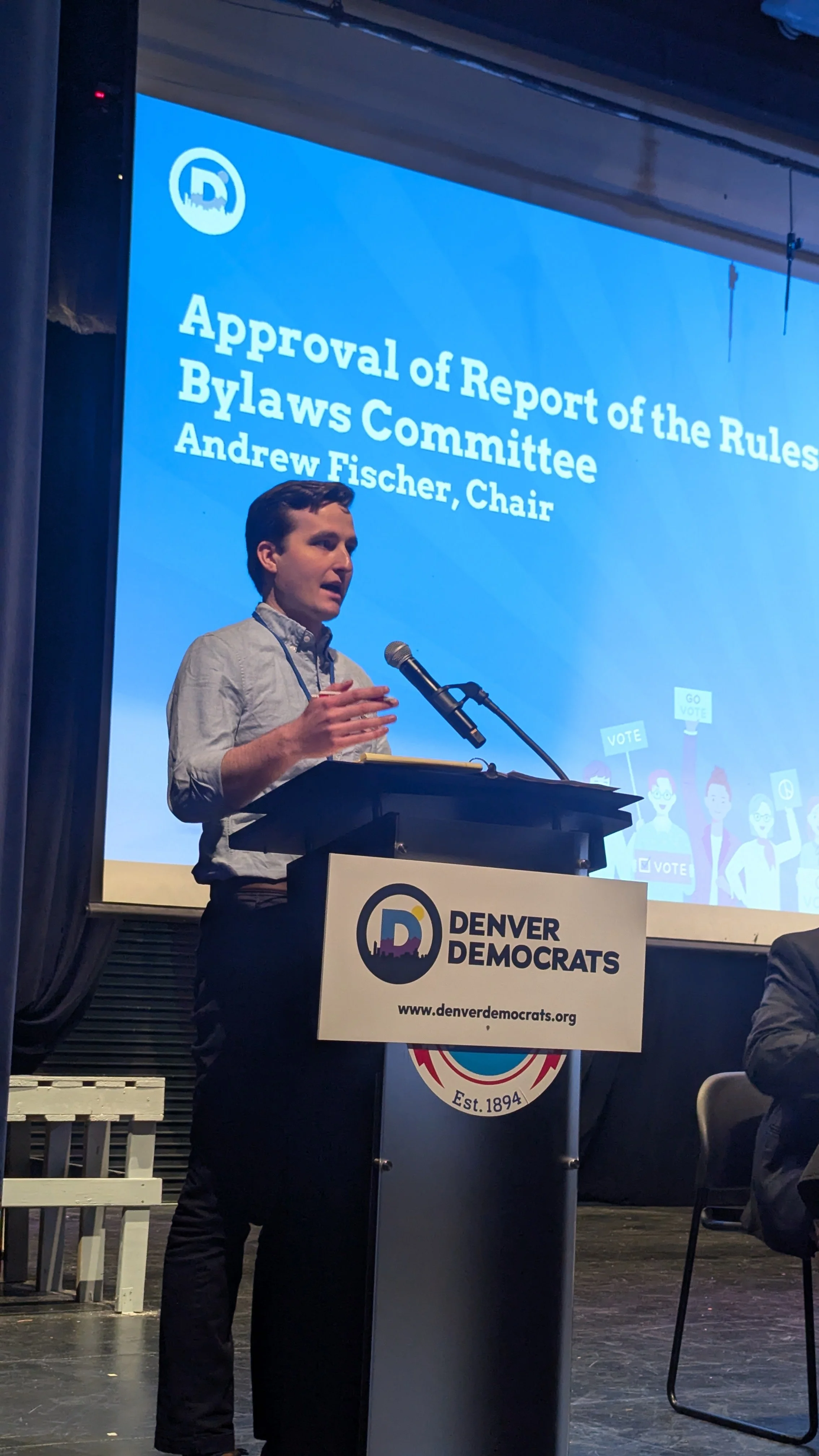 A man speaking at a podium with a Denver Democrats banner, during a meeting or event. Behind him, a large screen displays text about the approval of a report of the Rules Bylaws Committee, chaired by Andrew Fischer. The stage has a dark backdrop, and part of the audience can be seen.