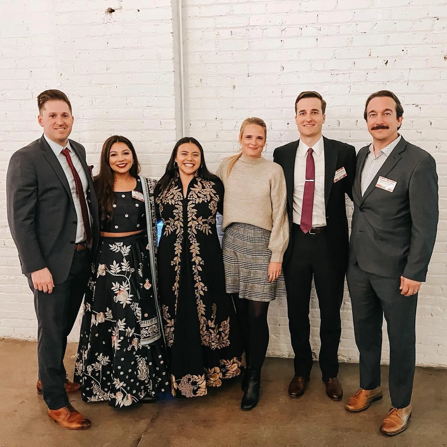 Six people standing against a white brick wall, some wearing name tags. They are dressed in formal and semi-formal attire, smiling for the photo.