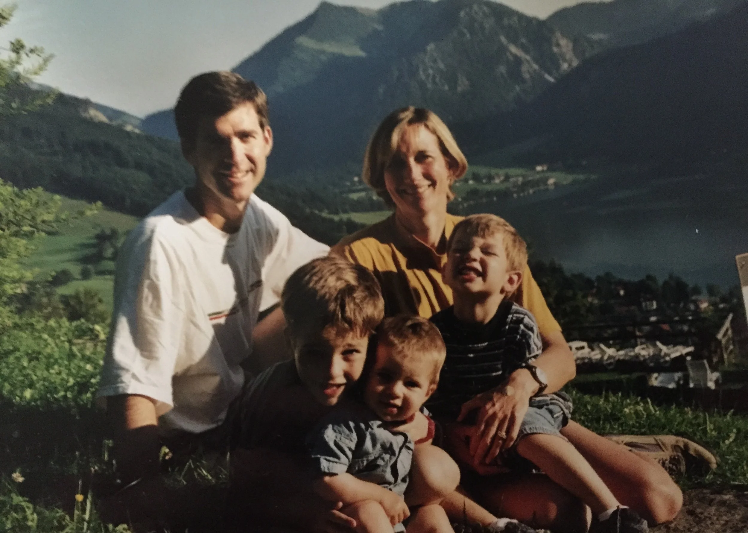 A group of children and two adults outdoors with mountains and green hills in the background, smiling and posing for a photo.