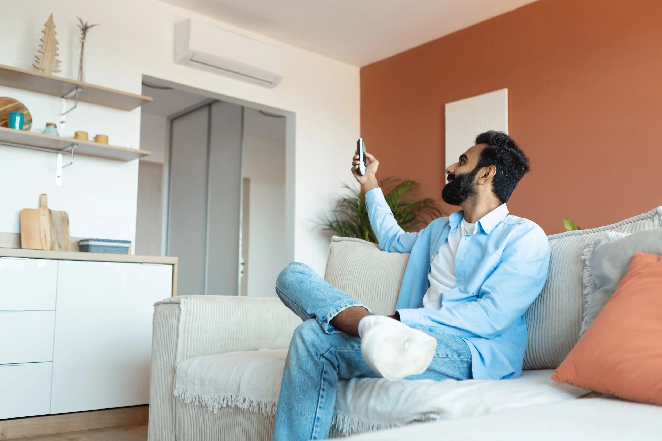 A man with dark hair, a beard, and wearing a light blue shirt, sitting on a beige sofa in a living room, holding a smartphone up to take a selfie, with a smile.