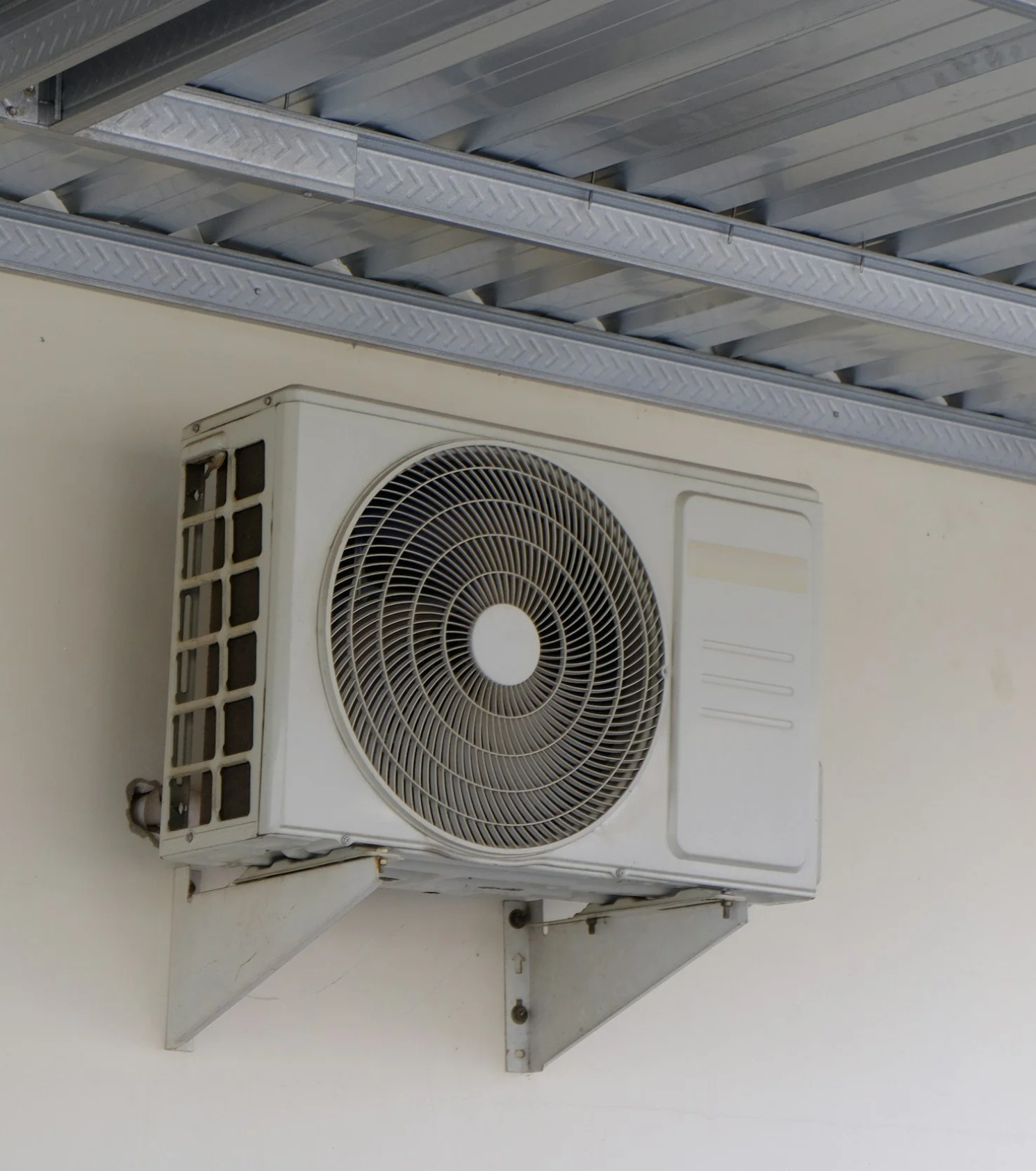 An air conditioning unit installed on a wall, with visible vents and a metal support bracket. Part of a metallic ceiling or awning is also visible above.