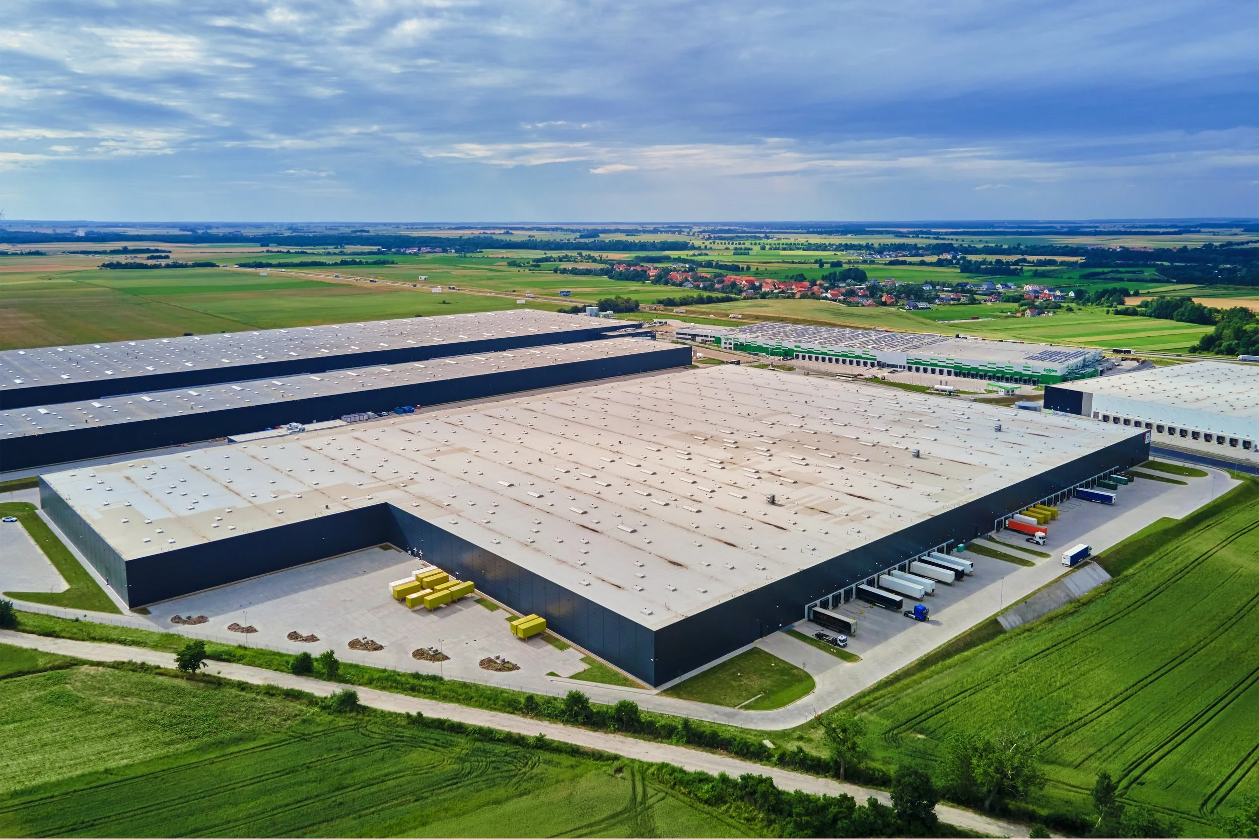 Aerial view of large industrial warehouse with loading docks, surrounded by green fields and a small town in the background under partly cloudy sky.