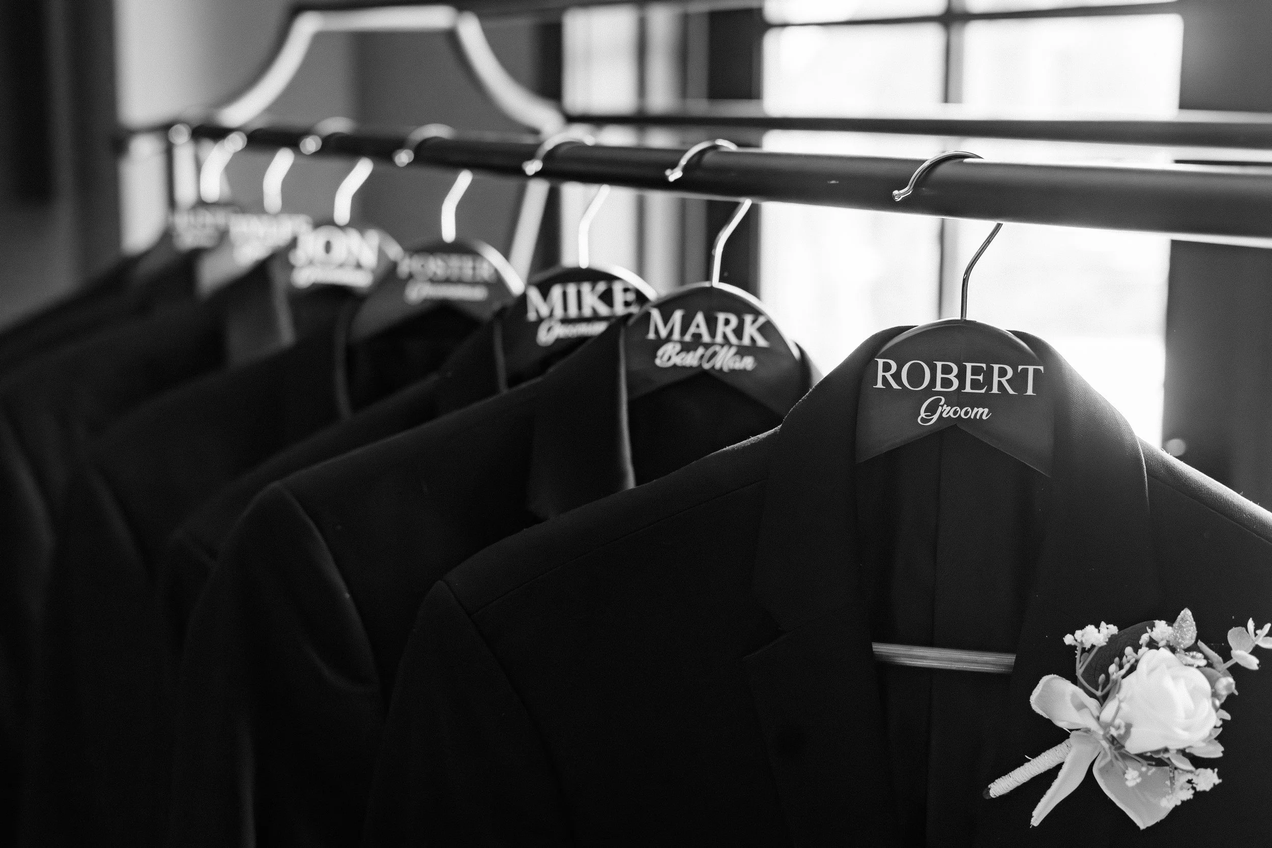Black and white photograph of suit jackets hanging on a rack, each with a personalized label indicating the person's name and role, such as groom or groomsmen. A bouquet of flowers is visible in the bottom right corner. Fourth Photography