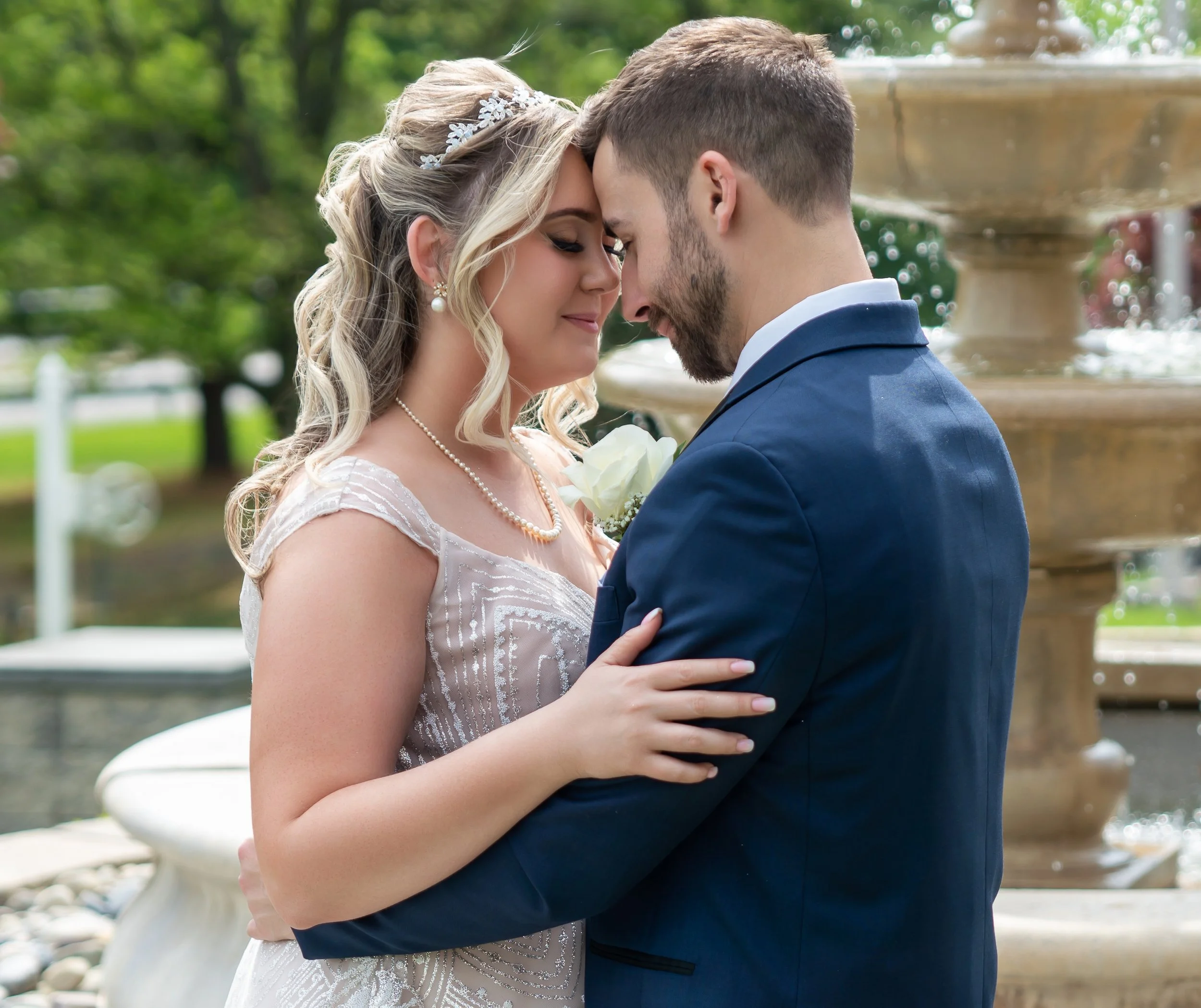 A bride and groom share a close moment with foreheads touching, outdoors near a fountain with green trees in the background. Fourth Photography