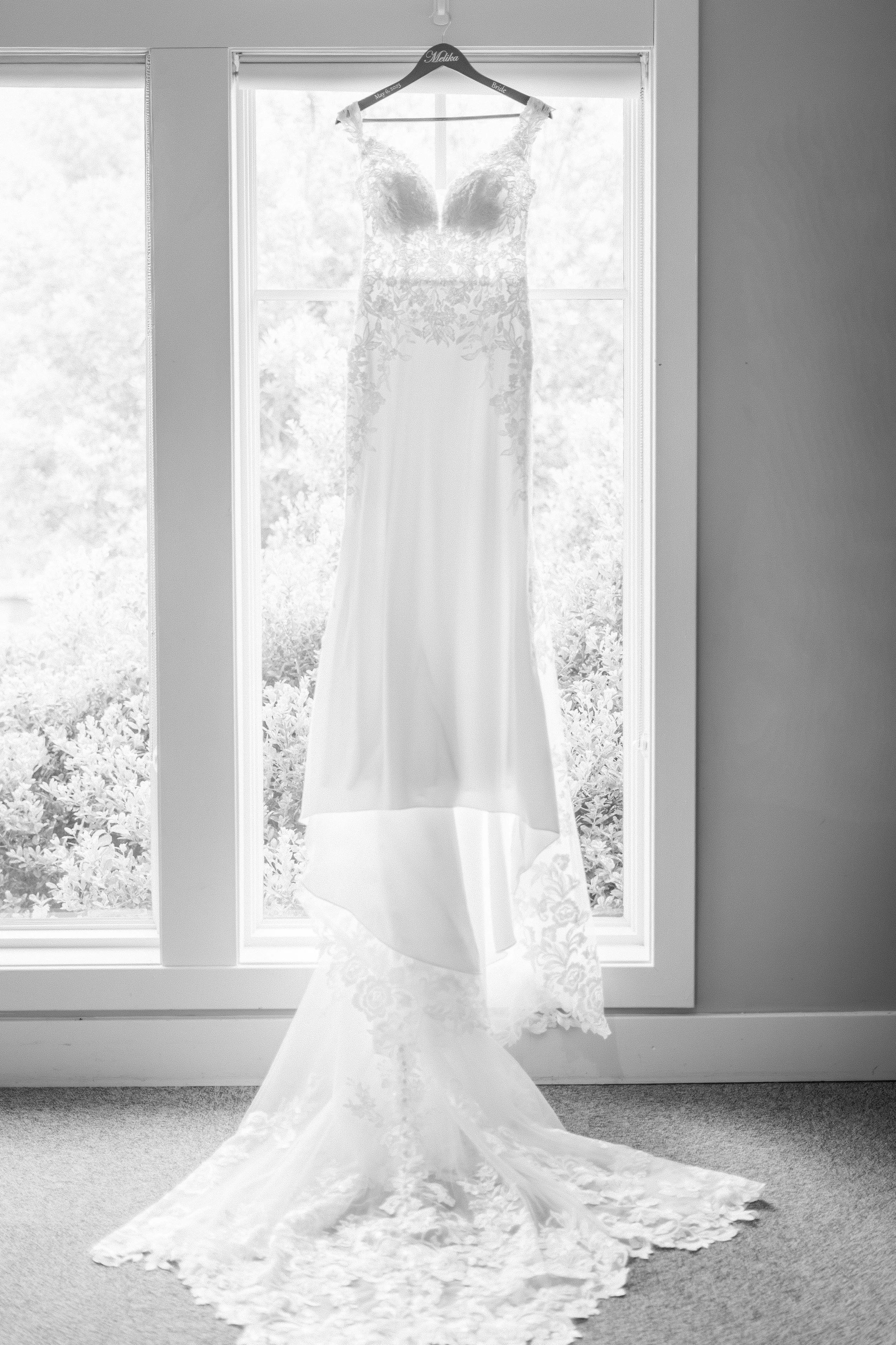 A wedding dress hanging on a black hanger with the names 'Malika' and 'Bride' and the date 'May 6, 2023', in front of a window. Fourth Photography