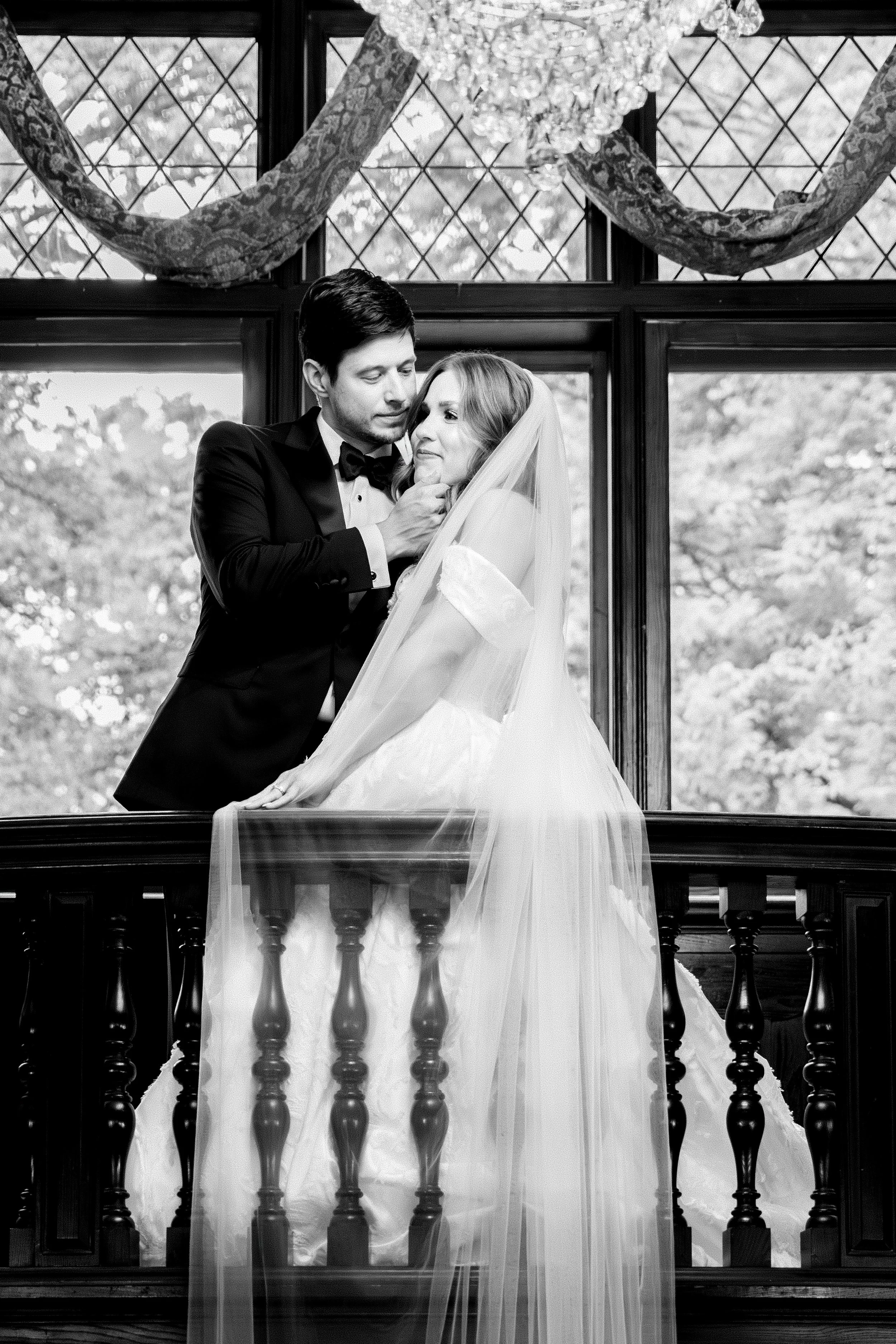 Black and white photo of a bride and groom standing on a balcony with balusters. The groom is dressed in a tuxedo and the bride is in a wedding dress with a veil. They are embracing each other with the groom gently touching the bride's face, sharing an intimate moment. The background shows windows with a landscape outside, and decorative curtains hang above.