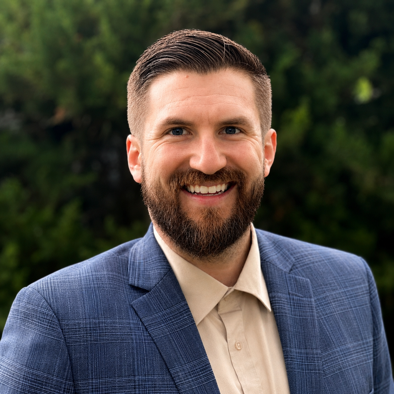 A smiling man with a beard and short dark hair, wearing a blue suit and a cream shirt, standing outdoors with green foliage in the background.