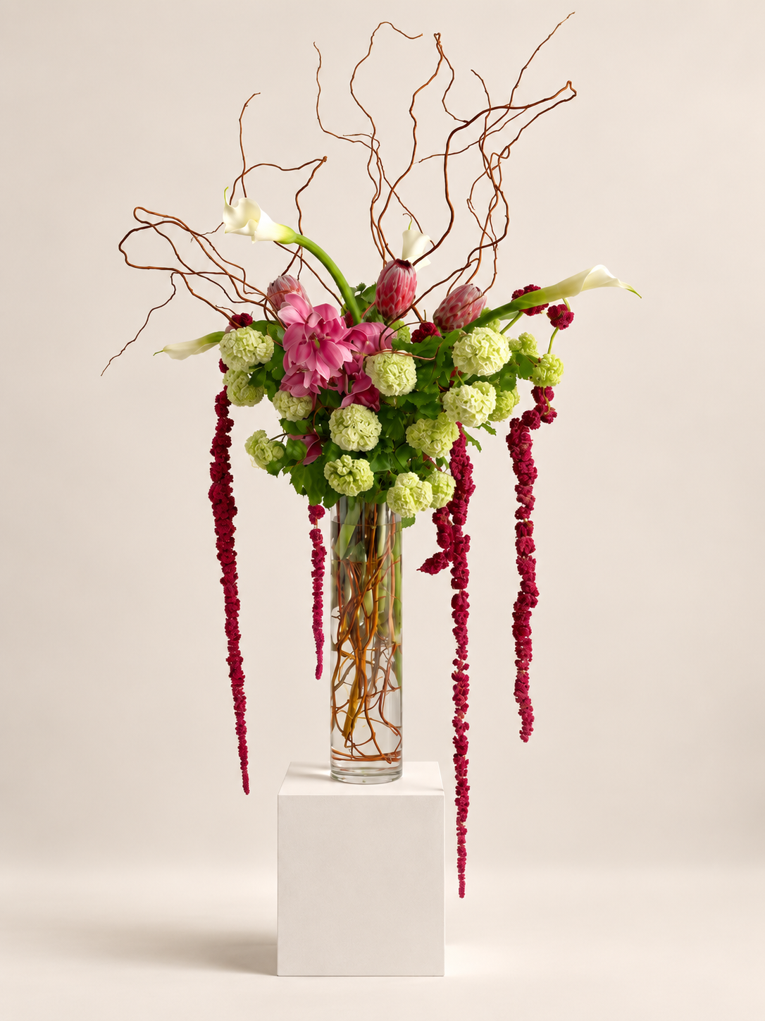Tall statement floral arrangement by Grove Floral Studios in Miami, featuring white calla lilies, pink flowers, green hydrangea, curly branches, and cascading burgundy amaranthus in a clear cylinder vase.