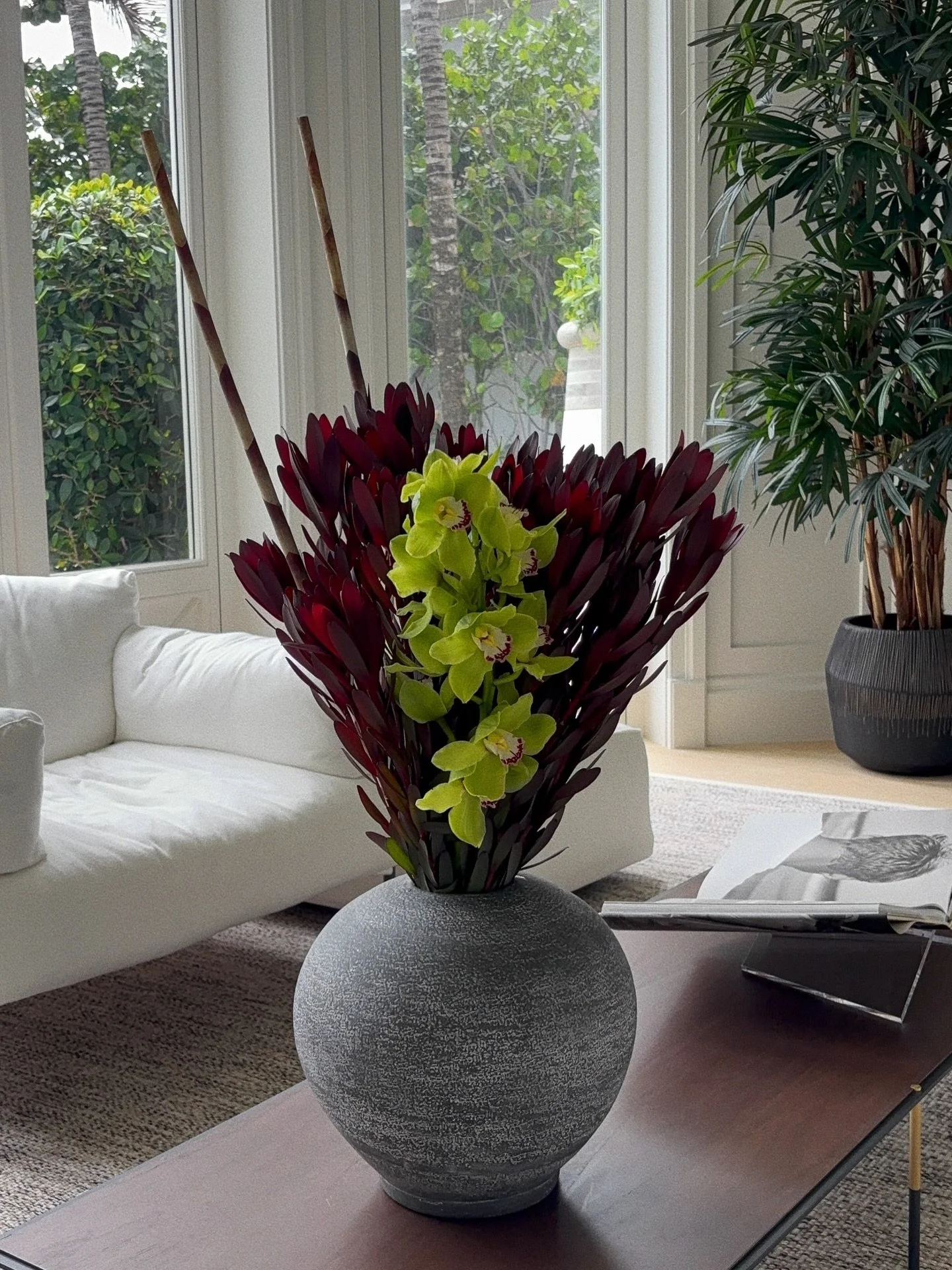 Dark burgundy leucadendron, green cymbidium orchids, and stripped bare branches in a spherical stone vase. Dramatic without trying.
Available for your home &mdash; DM to book &middot;
