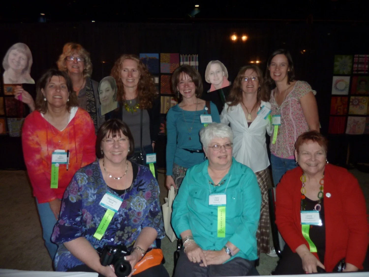 The Twelve by Twelve art quilt group photographed together at the International Quilt Festival in Houston, with face cutouts representing absent members.