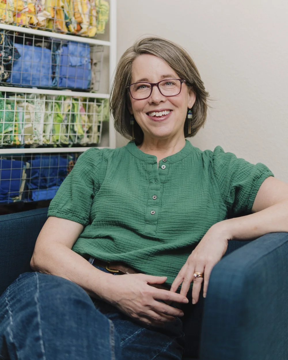 Artist and author Deborah Boschert wearing a green blouse, sitting on a dark couch in front of a shelf filled with colorful yarn.