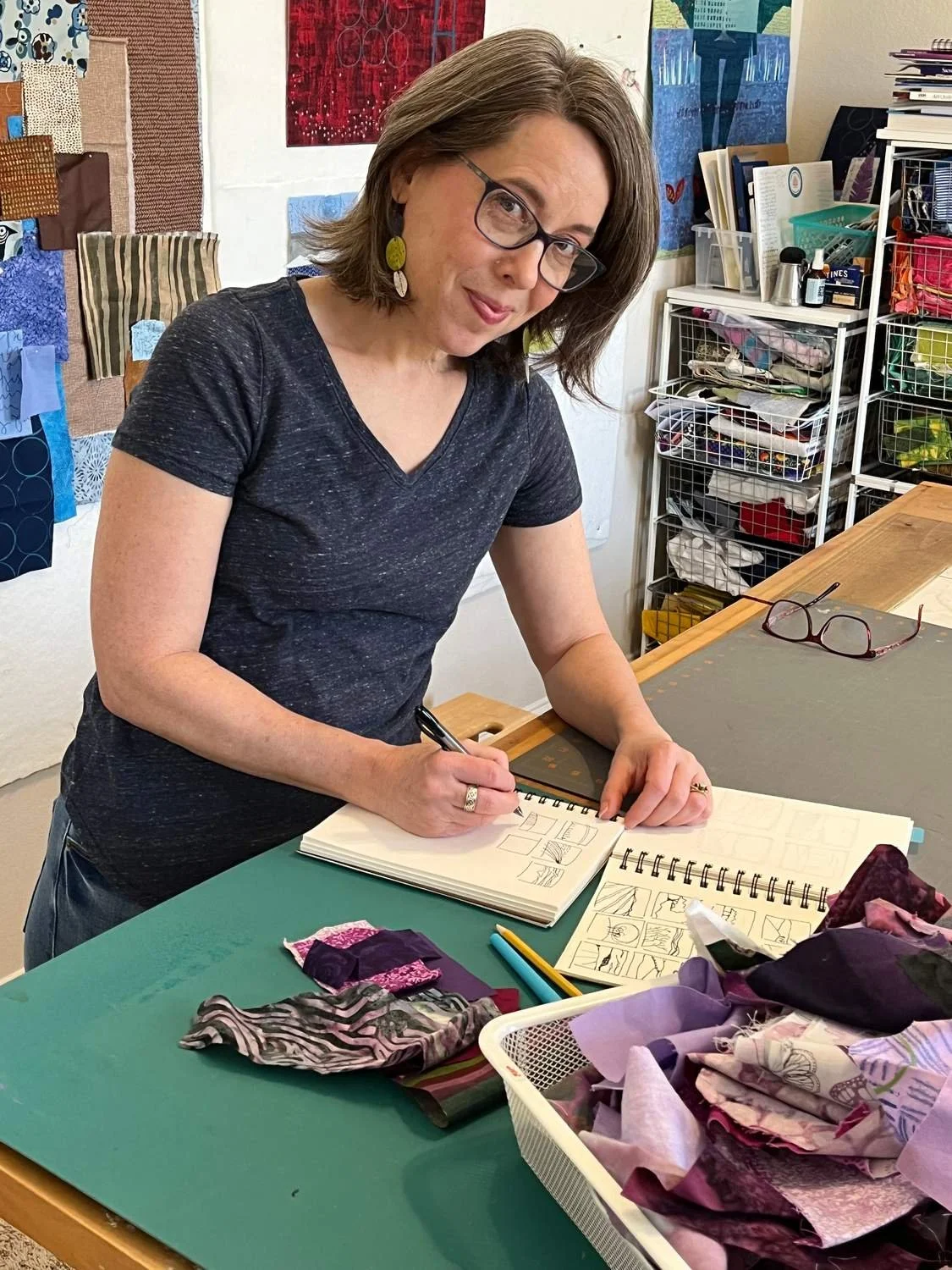 A woman with glasses and earrings is standing at a work table, drawing in a sketchbook. She is in an art studio with fabric and artwork on the walls and shelves filled with supplies and fabric in the background.