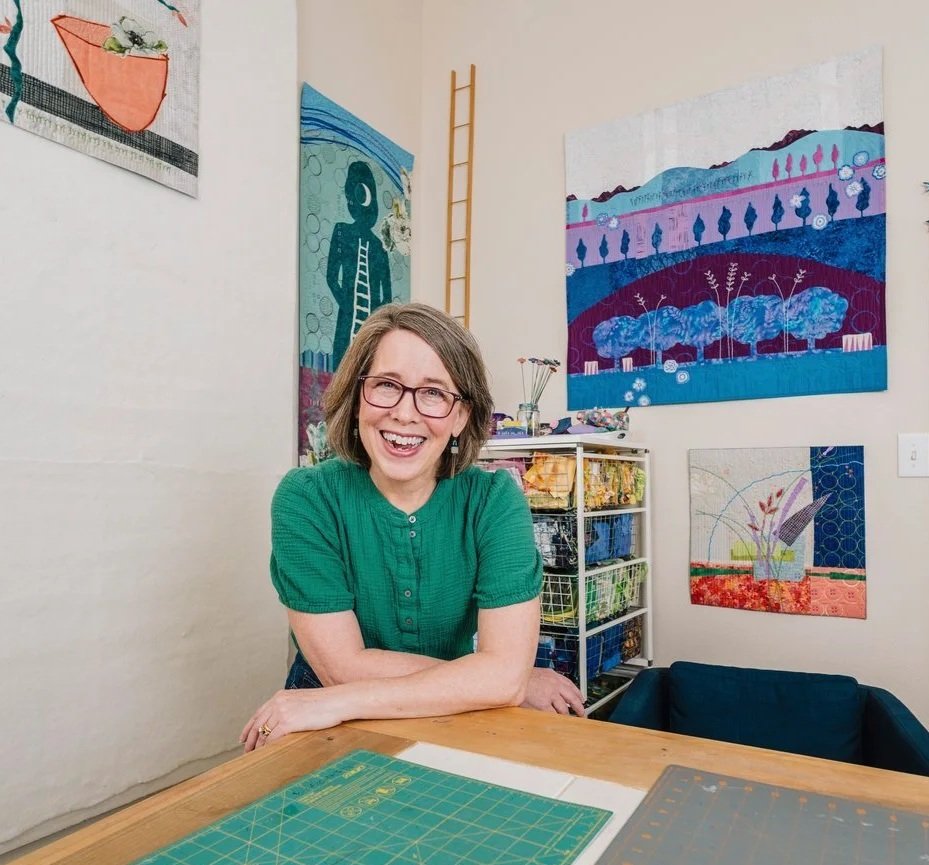 A woman with glasses and wearing a green shirt smiling at a wooden table in her art studio, with colorful paintings on the walls and art supplies on a shelf behind her.