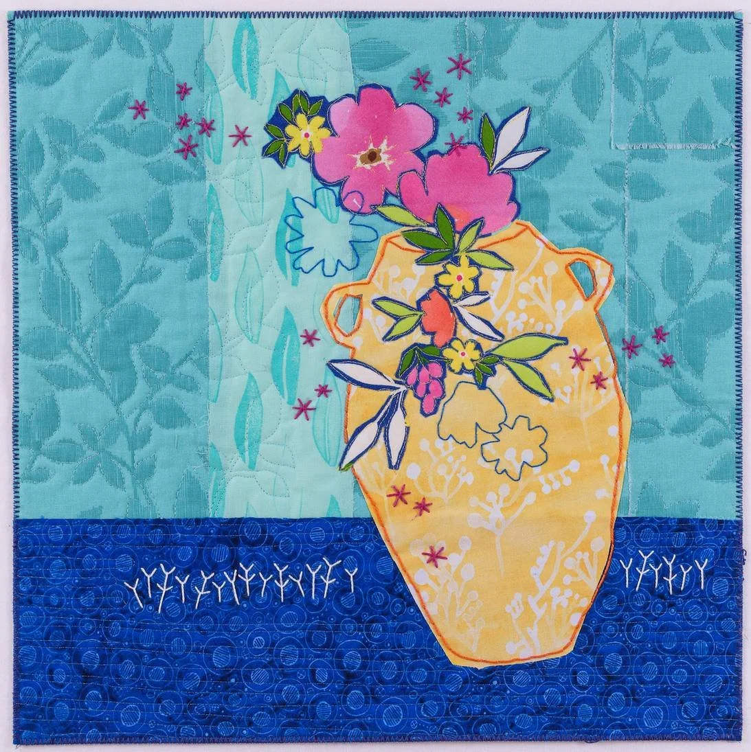 A colorful quilt with a flower vase design, featuring pink, yellow, and purple flowers on a blue background with leaf patterns and embroidered details.