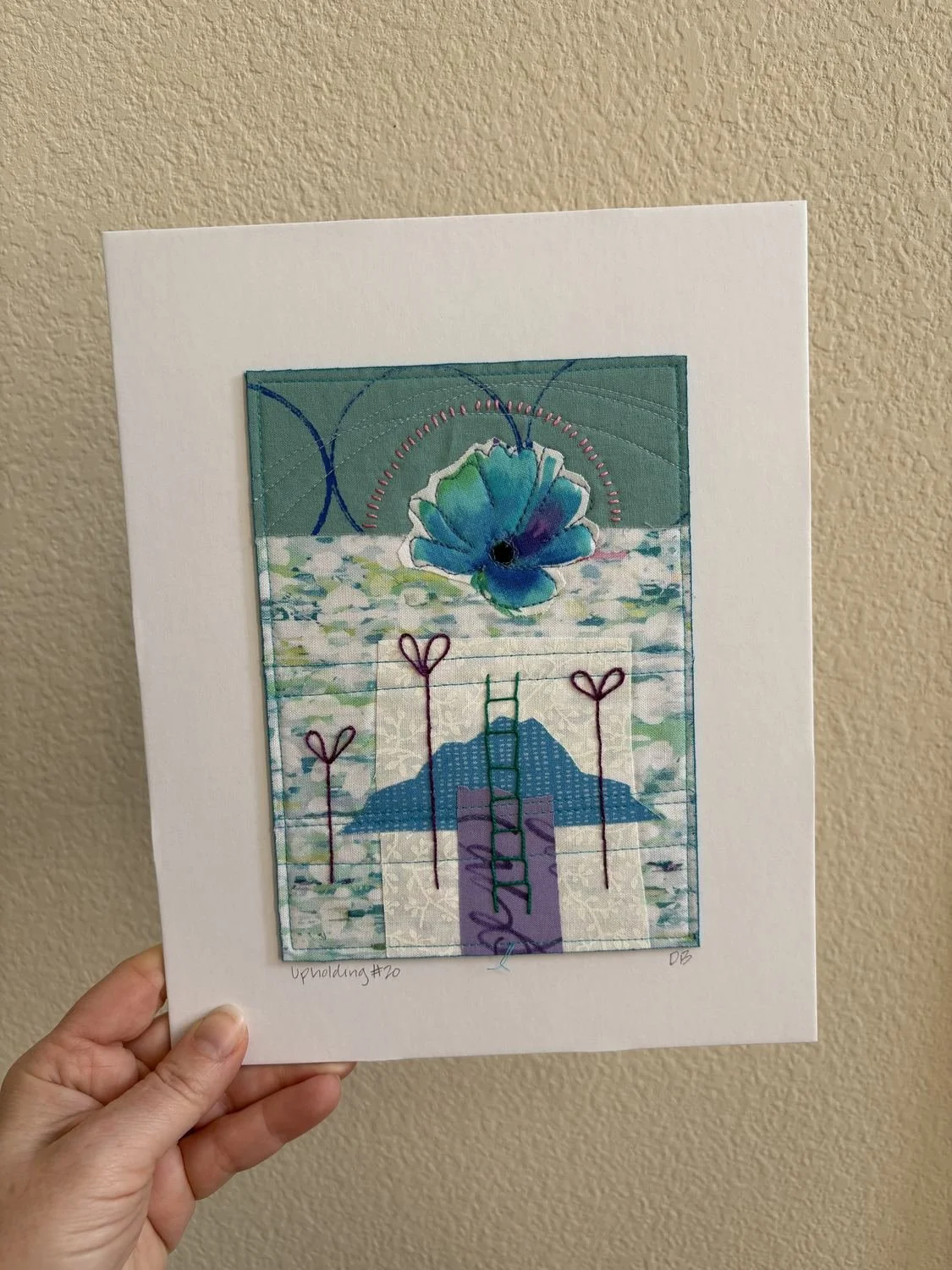 A quilting art piece featuring a blue flower with green and purple accents, stitched with thread, set against a background with mountains, hearts, and water patterns, mounted on a white page.