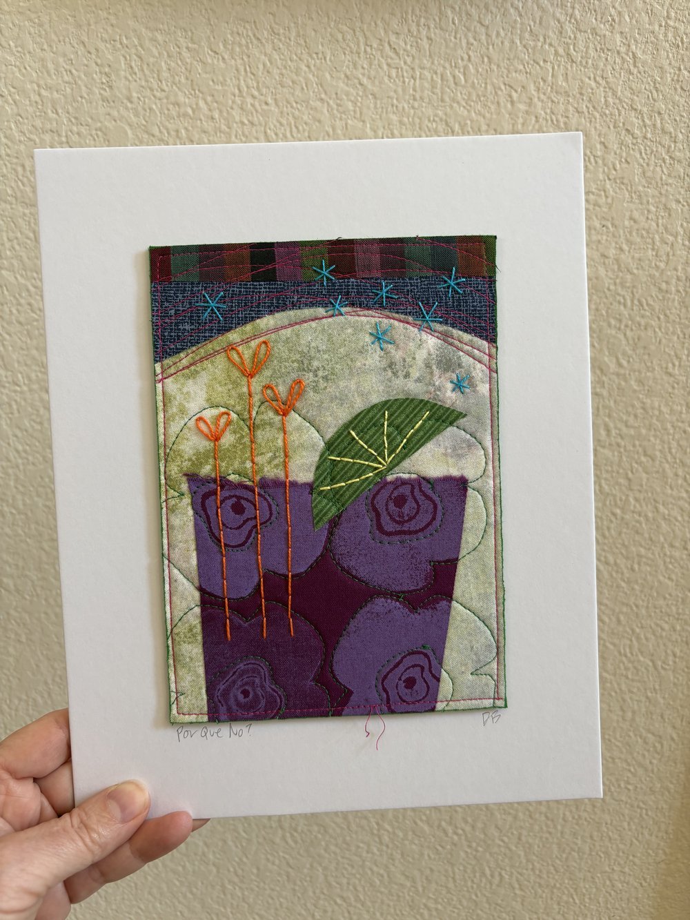 A detailed fabric art piece with embroidered floral and star designs, mounted on white background paper.