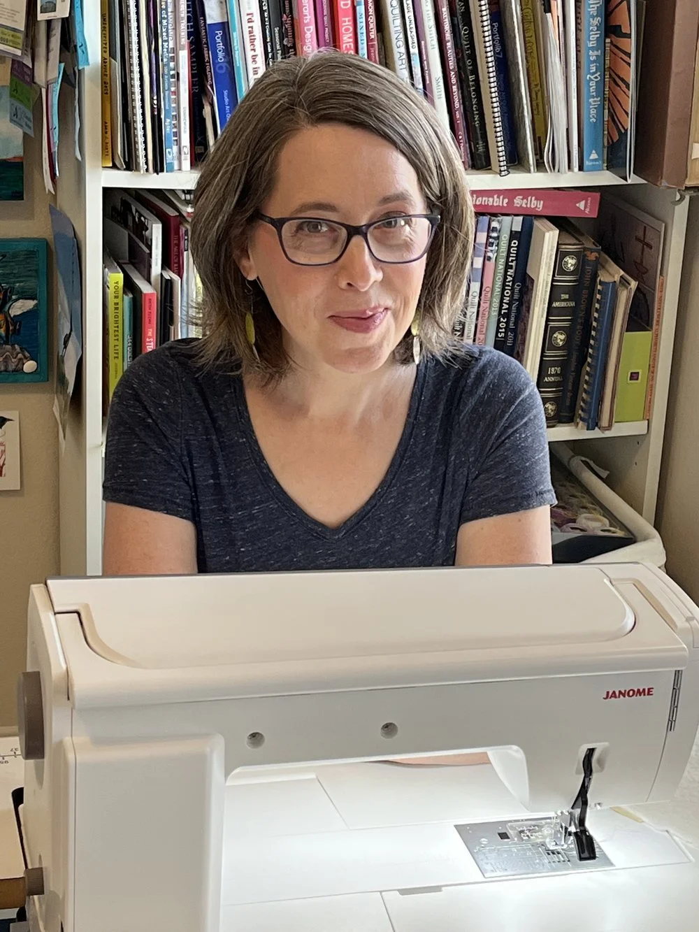 A woman with glasses and shoulder-length hair sitting in front of a sewing machine, with a bookshelf filled with books in the background.