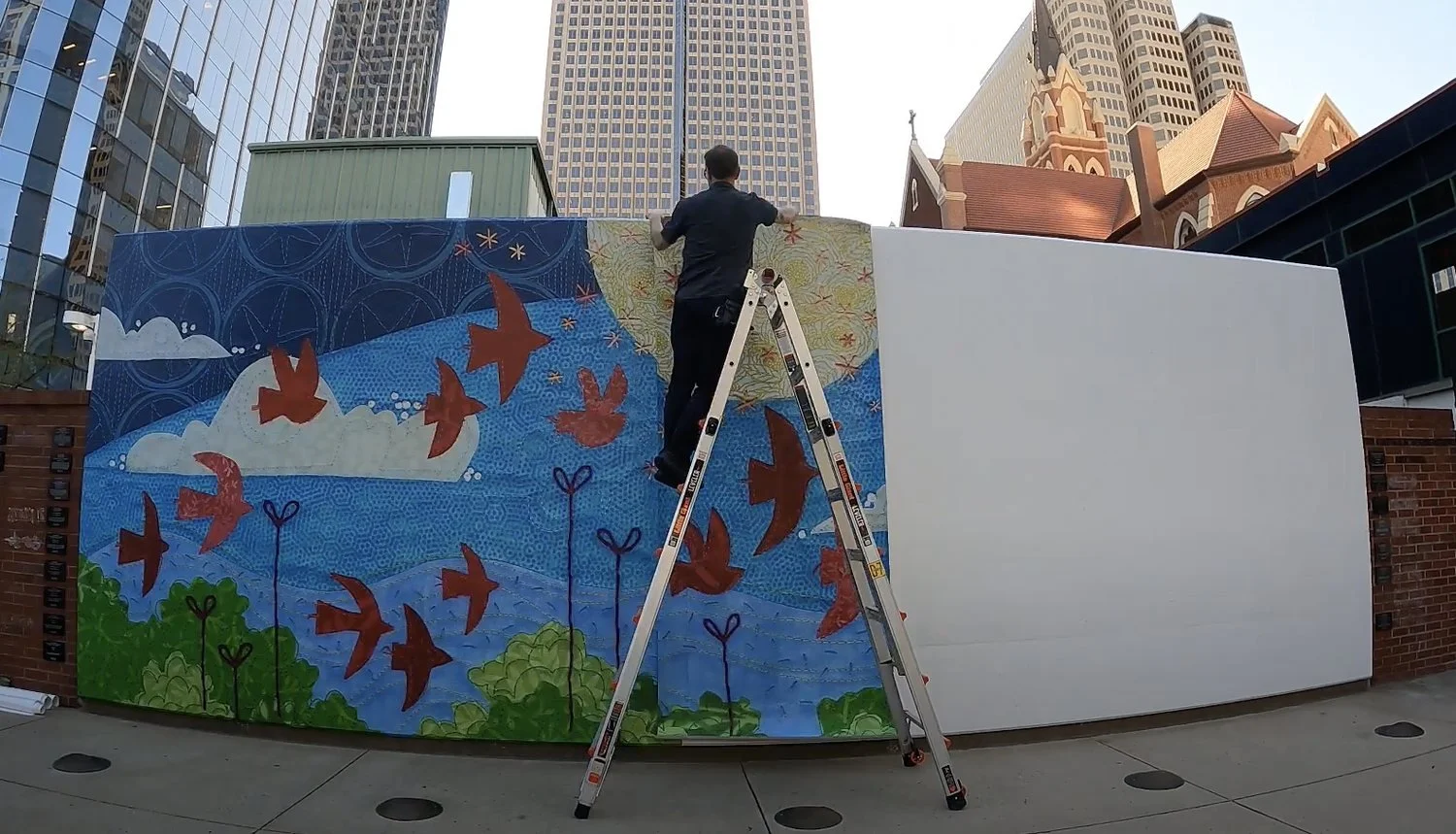 Worker on a ladder installing panels of the Looking Up mural on an outdoor downtown wall.
