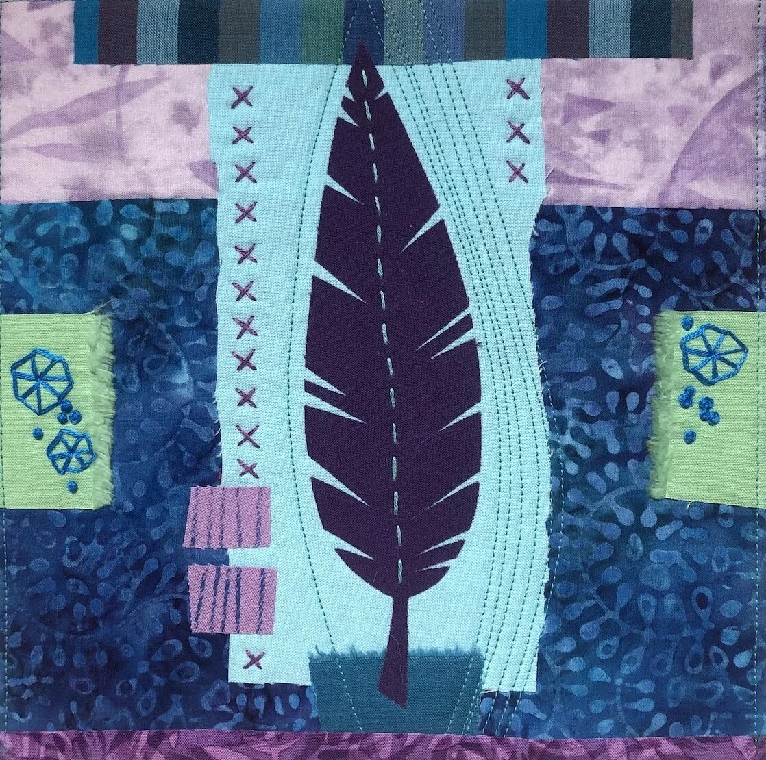 Close-up of a quilt featuring a central design of a stylized dark blue bird feather on a turquoise background with various patterned fabric patches surrounding it.