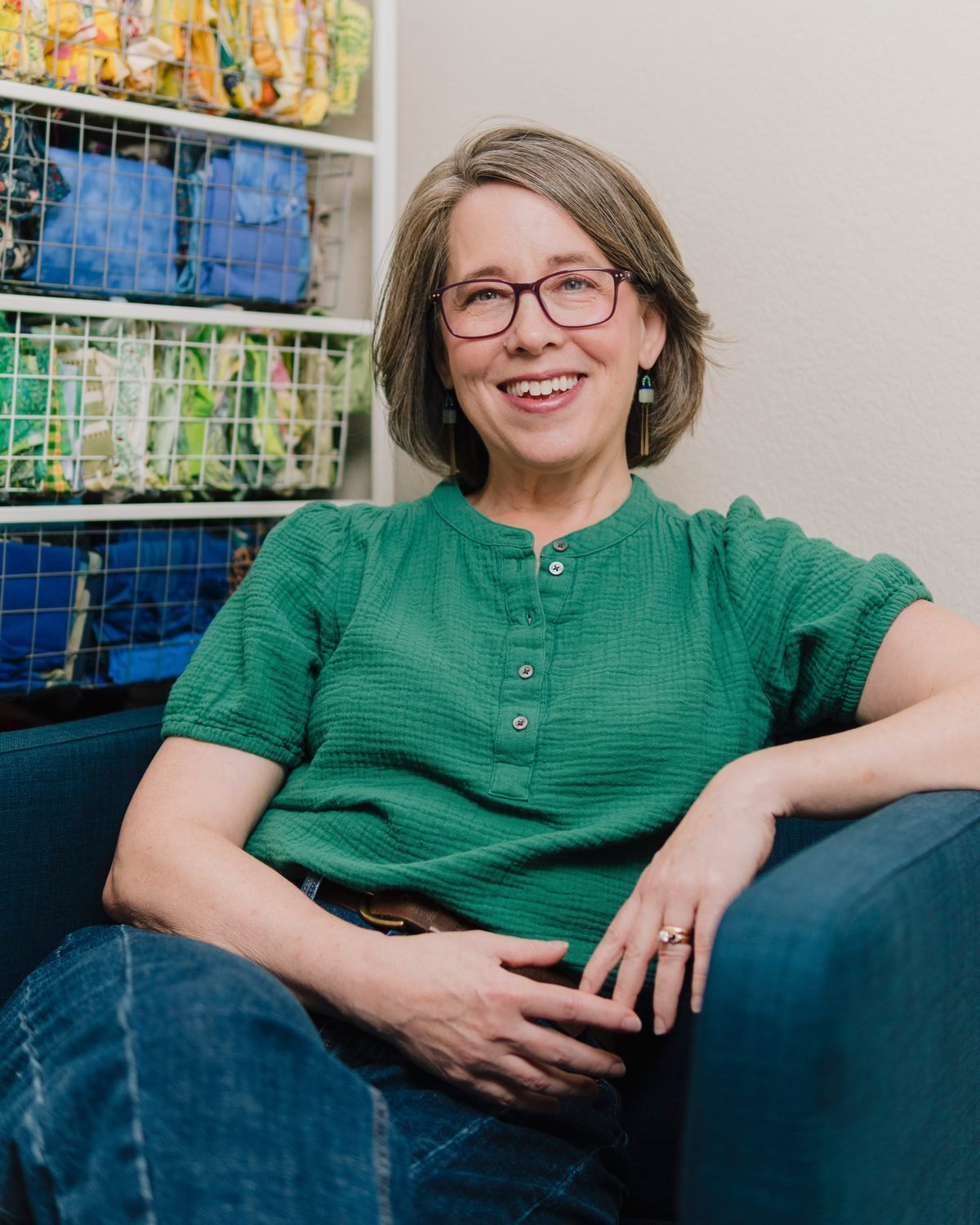 A woman with shoulder-length gray hair, glasses, and earrings is smiling and taking a selfie while sitting on a dark blue couch. She is wearing a green, short-sleeved, buttoned blouse and dark jeans. Behind her, there are shelves filled with colorful yarn and yarn balls.