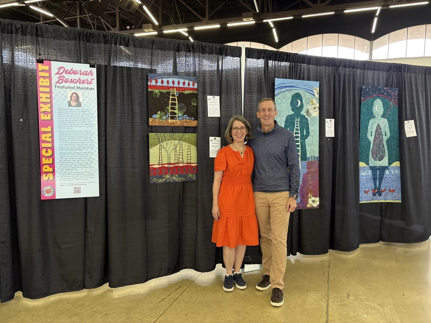 Deborah Boschert standing with a visitor in her Dallas Quilt Show booth beside her ladder-themed figurative art quilts.