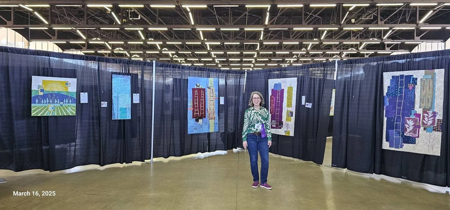 Deborah Boschert standing in her special exhibit booth at the Dallas Quilt Show, surrounded by her art quilts on draped display walls.