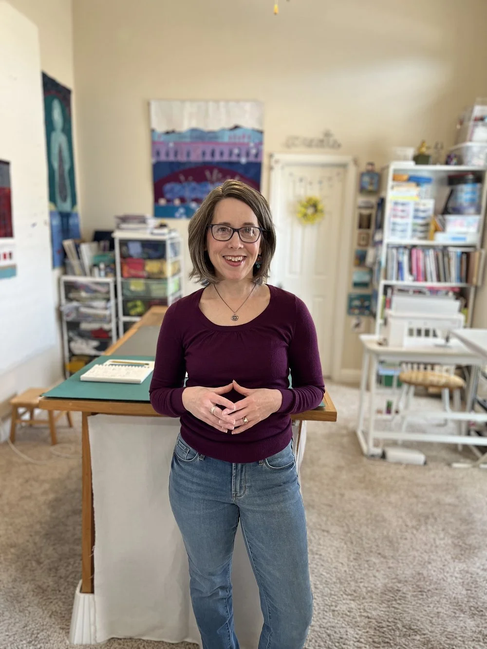 Woman with glasses, wearing a burgundy sweater and blue jeans, standing in a craft room or classroom with shelves and art supplies in the background.