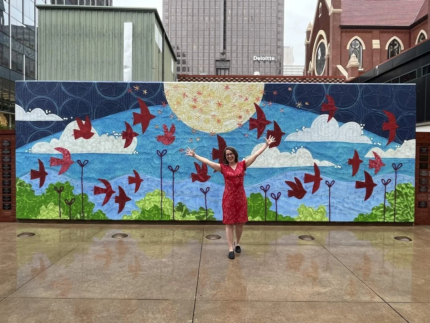 A woman in a red dress stands in front of a colorful mural with her arms raised, smiling. The mural features a large yellow sun, blue sky with clouds, green hills, and numerous red birds flying across the sky, in a stylized, abstract pattern.