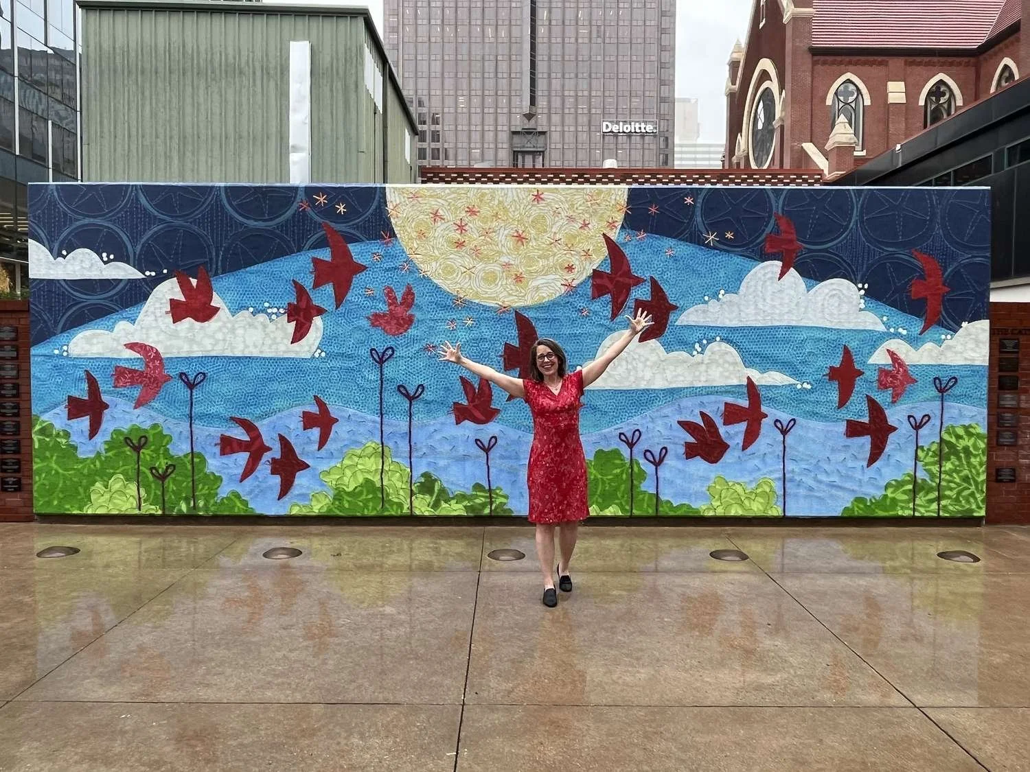Deborah Boschert standing with arms raised in front of her large outdoor "Looking Up" mural in downtown Dallas.