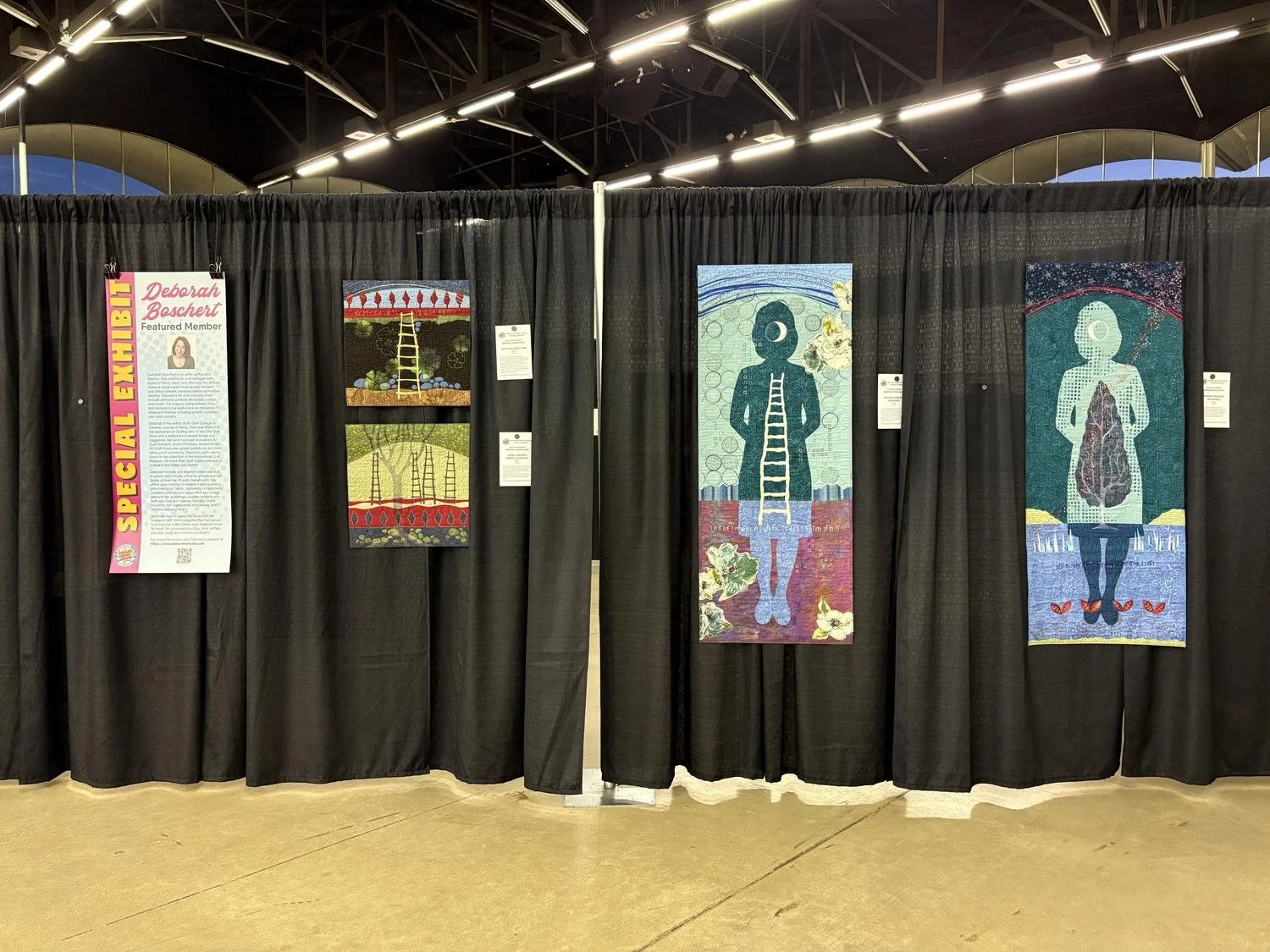 Front panel of Deborah Boschert's Dallas Quilt Show booth showing her Featured Member banner and three figurative art quilts.