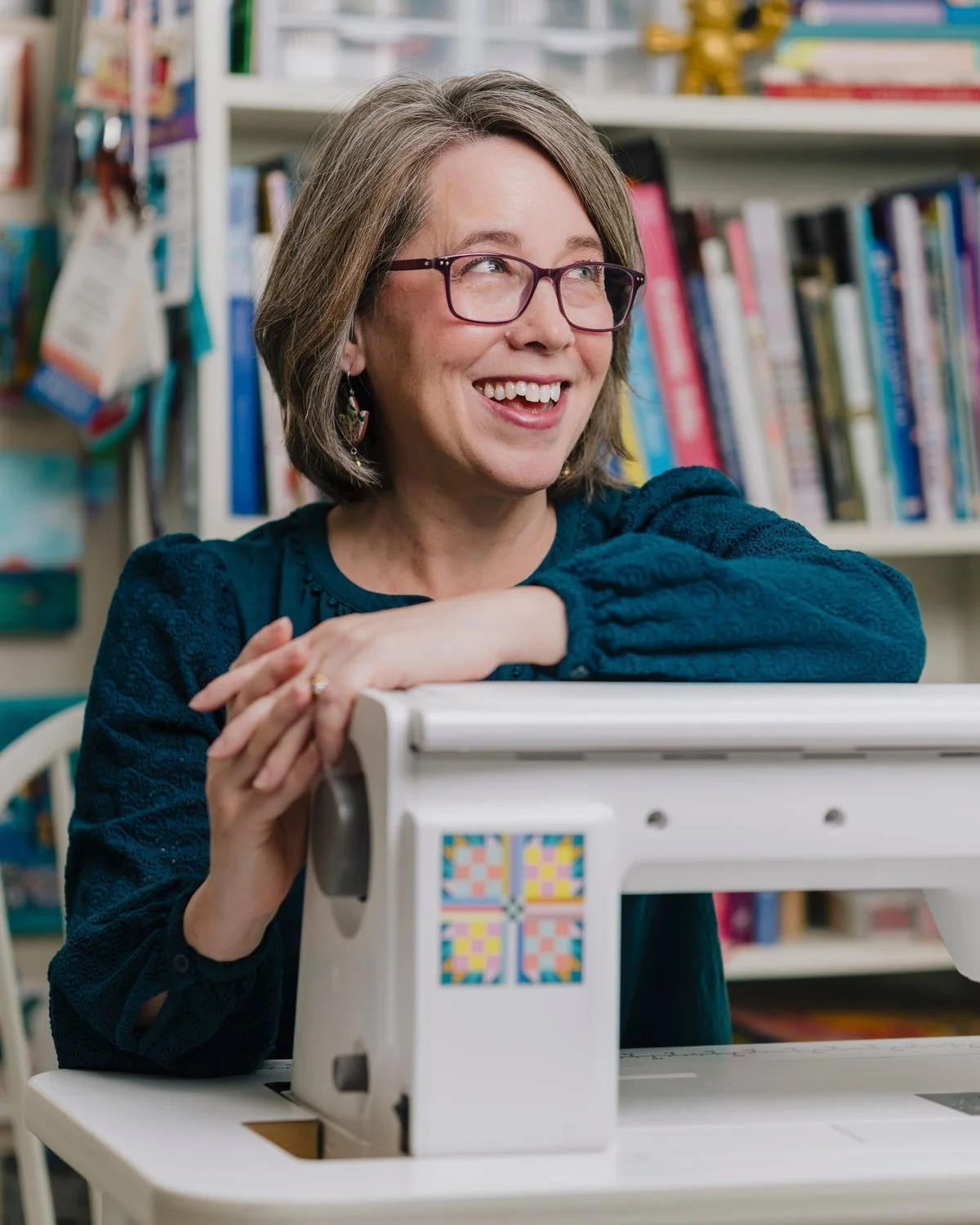 A woman with glasses smiling and resting her arms on a sewing machine, in a library or bookstore setting with books on shelves in the background.