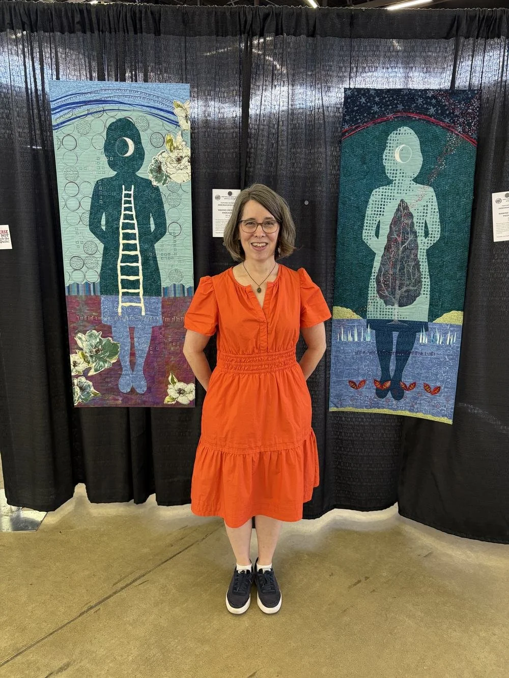Deborah Boschert in an orange dress standing between two of her self-portrait figurative art quilts at the Dallas Quilt Show.