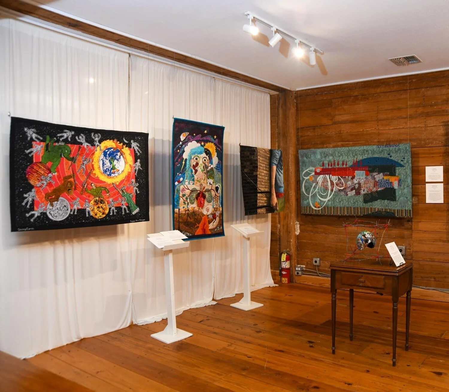 Art exhibit featuring three textile art pieces hanging on a white curtain and a wooden wall. The artworks are colorful and abstract, displayed with informational placards below each. A small table with a globe sculpture and a wire frame structure is positioned nearby, with a fire extinguisher on the floor and a window with blinds.