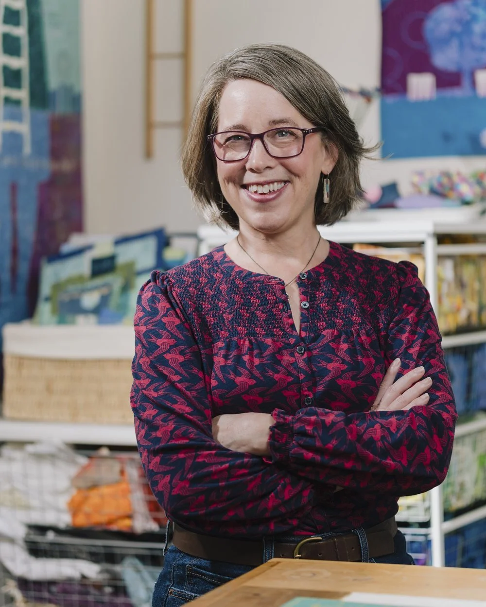 A woman with short gray hair, glasses, and a patterned red and purple blouse standing with arms crossed in a colorful classroom or art room.