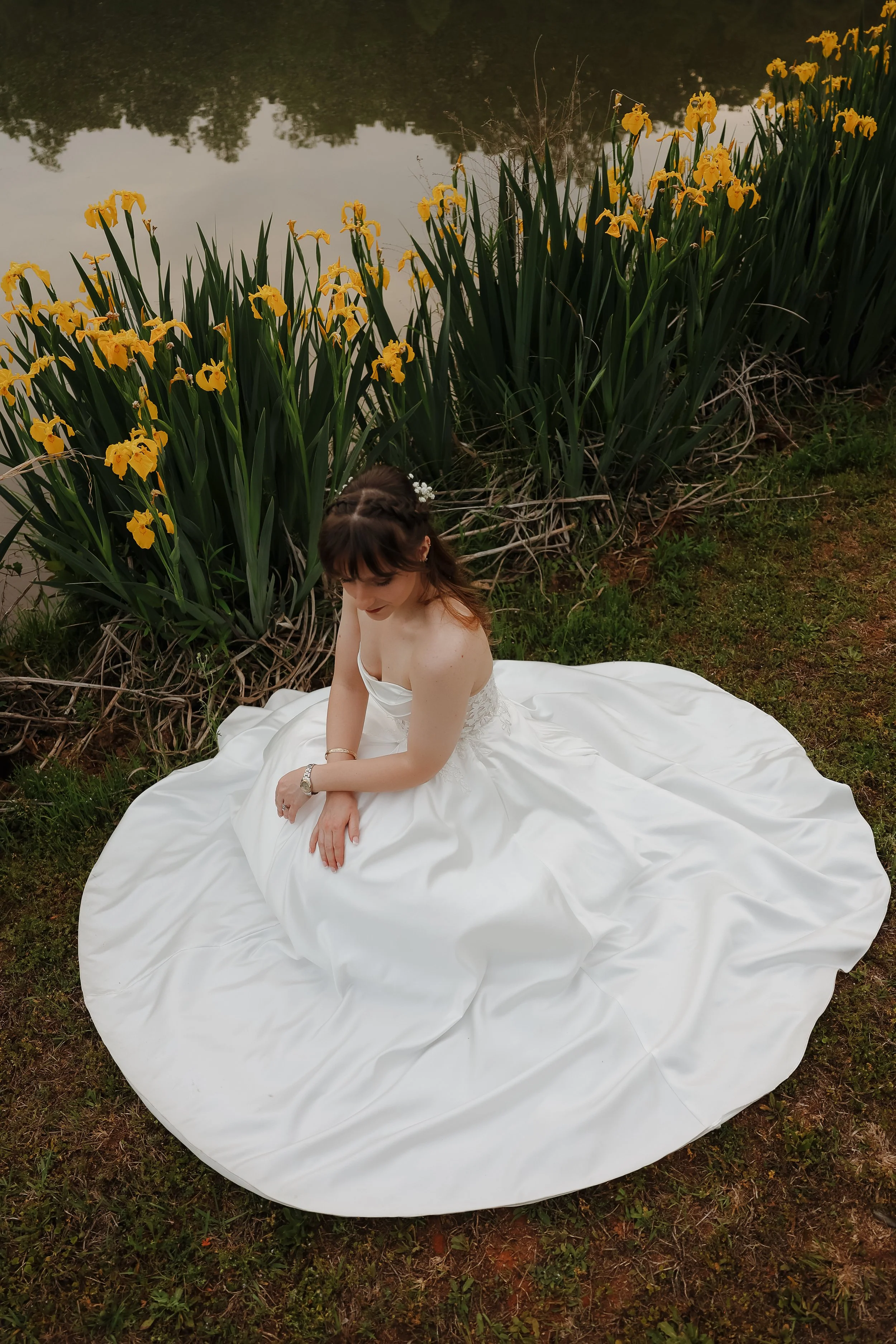 A woman in a white dress sitting on the grass near yellow irises by a lake, with trees reflected in the water in the background.