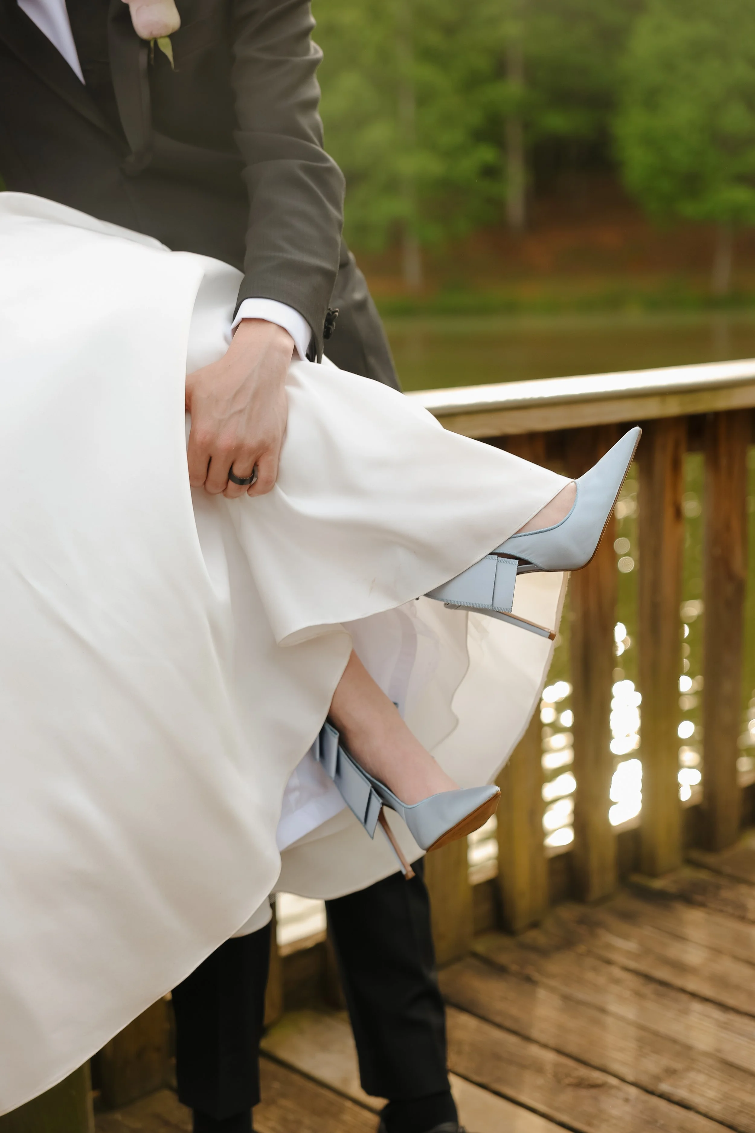 Close-up of a wedding ceremony scene with a groom lifting the bride's dress on a wooden deck by a lake with trees.
