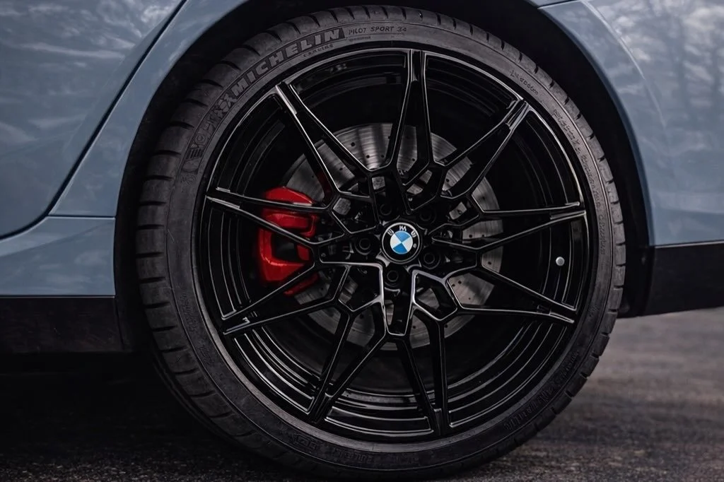 Close-up of a BMW car wheel with black rim, red brake caliper, and Michelin Pilot Sport tire, on a dark asphalt road.