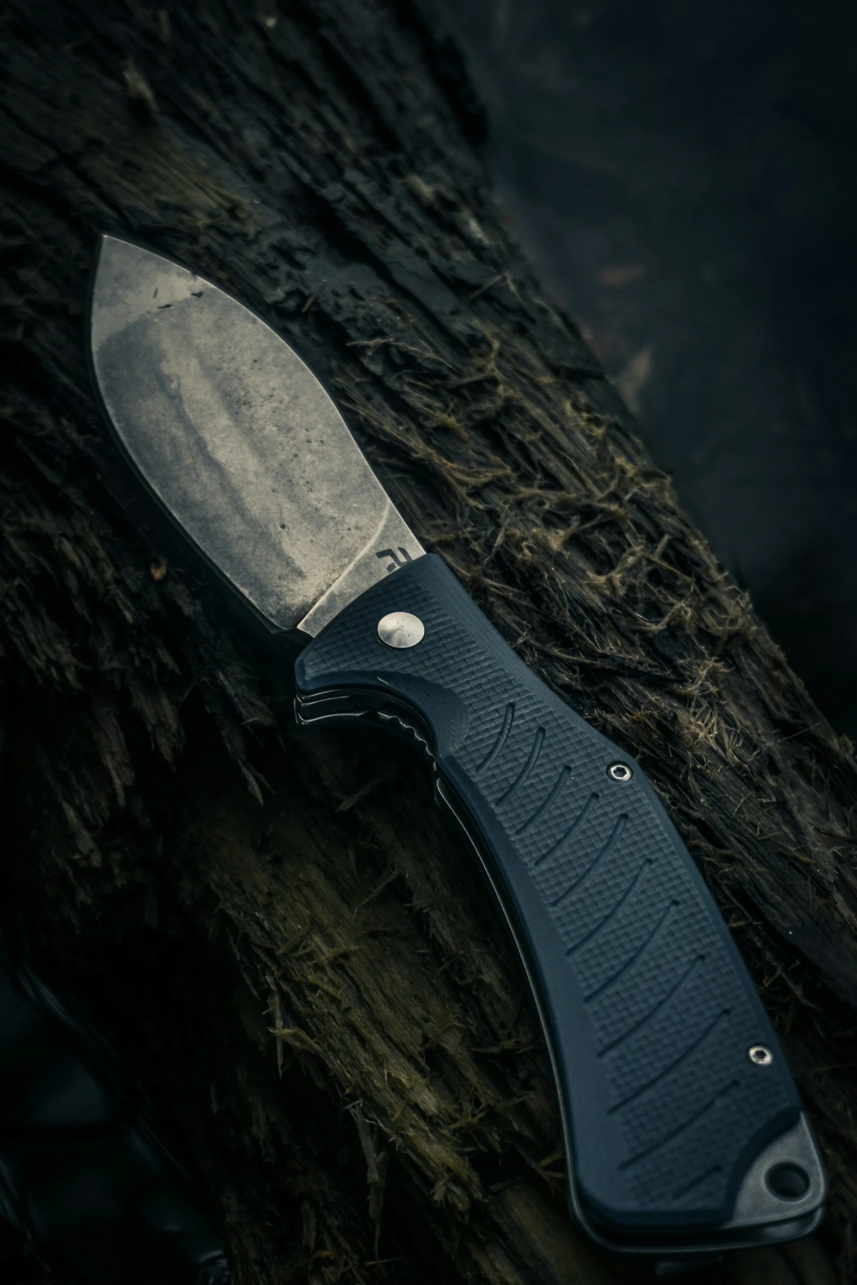 Close-up of a fixed-blade hunting or survival knife with a textured gray handle resting on a rough, moss-covered log in a dark outdoor setting.