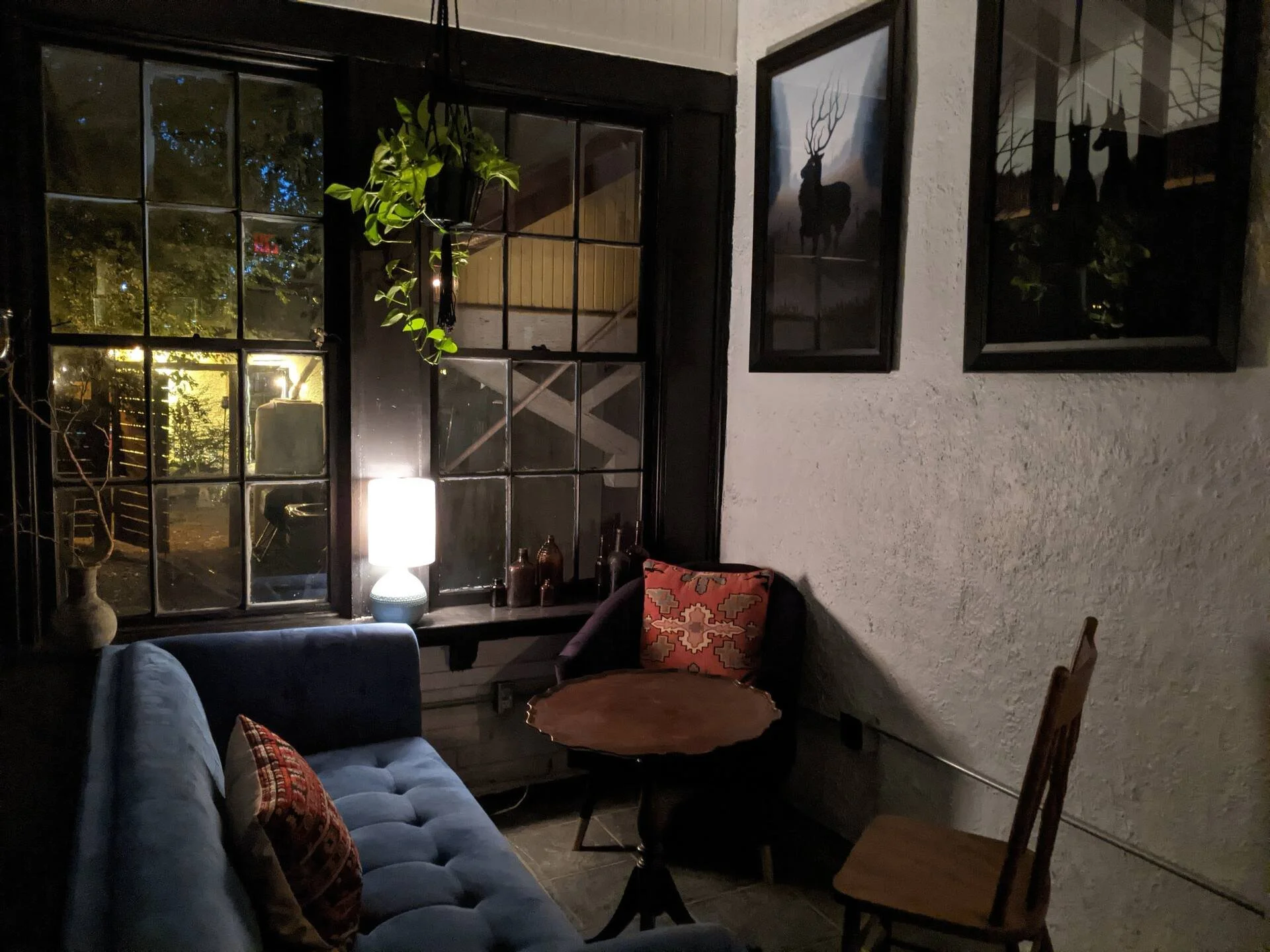 Living room with a large window, blue couch with colorful cushions, a side table with a lamp, two framed pictures of deer with antlers on the wall, and a wooden dining chair.