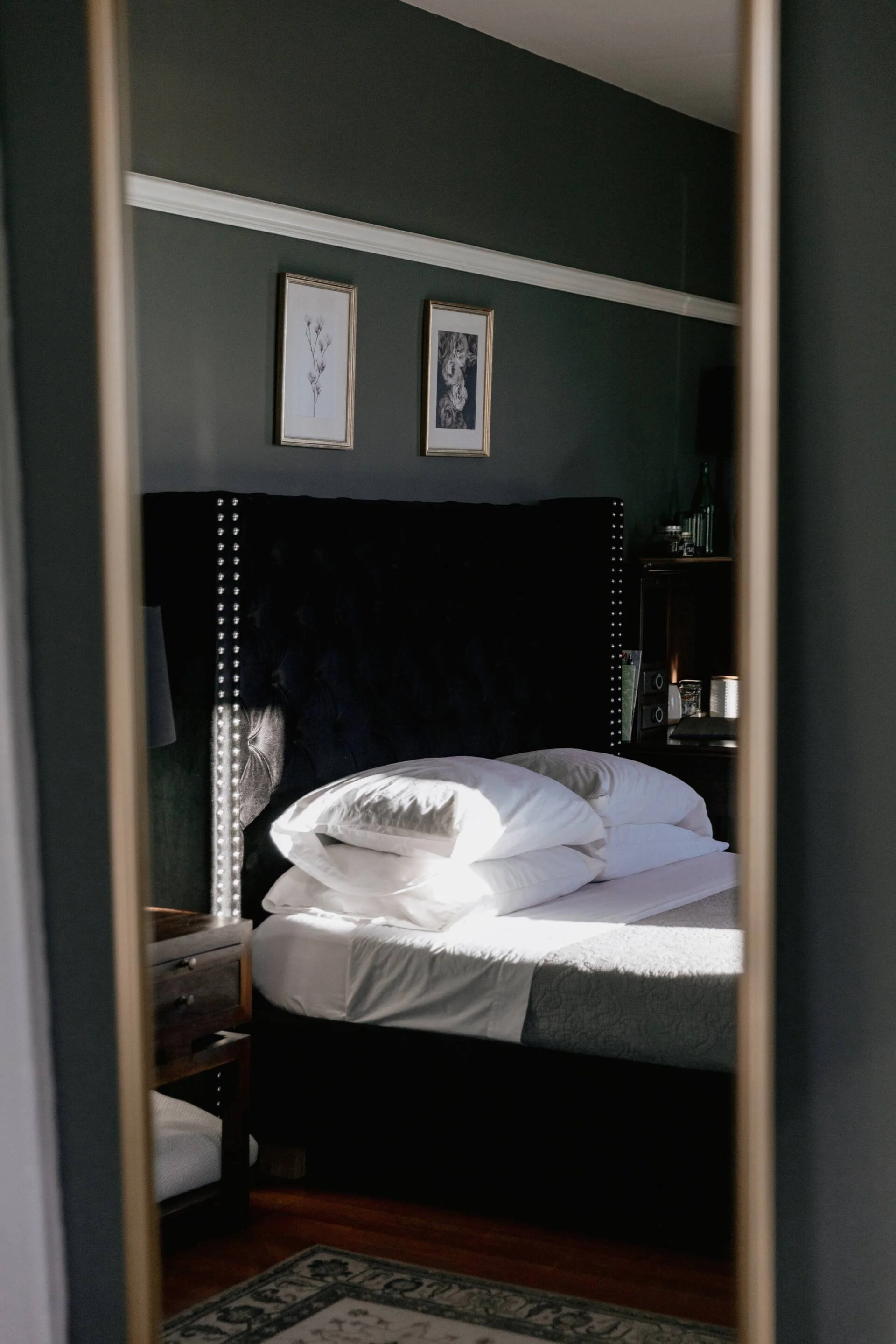 A bedroom with a dark green wall, framed artwork, a black tufted headboard with nailhead trim, a bed with white pillows and sheets, a bedside table with a lamp and decorations, and a patterned rug on the wooden floor, viewed through a mirror.