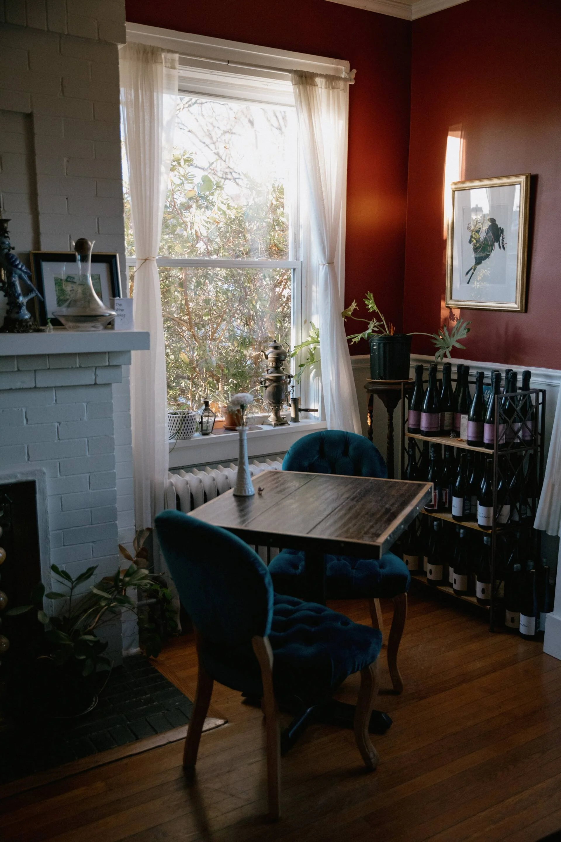 A cozy dining area with a wooden table and plush blue chairs. The room features a white brick wall, a window with white curtains letting in natural light, and a red-painted wall. There are decorative items on a shelf, and a wine rack with bottles.