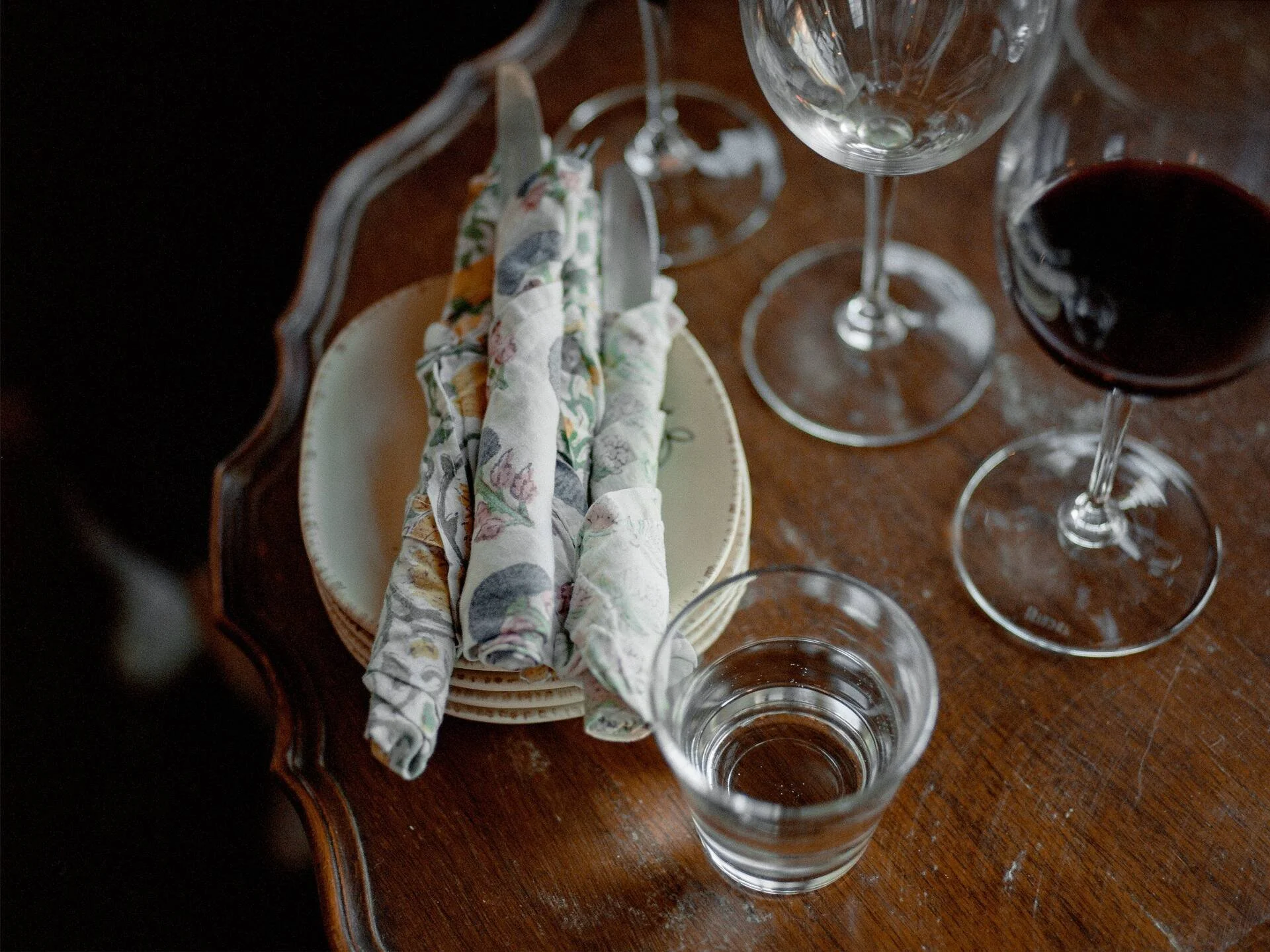 Empty wine glass and a partially filled glass of red wine on a wooden table next to a decorative ceramic basket with fabric containing utensils.